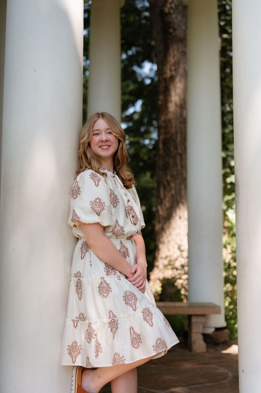 Teen girl in cream paisley dress smiles while leaning against white columns for senior portraits.