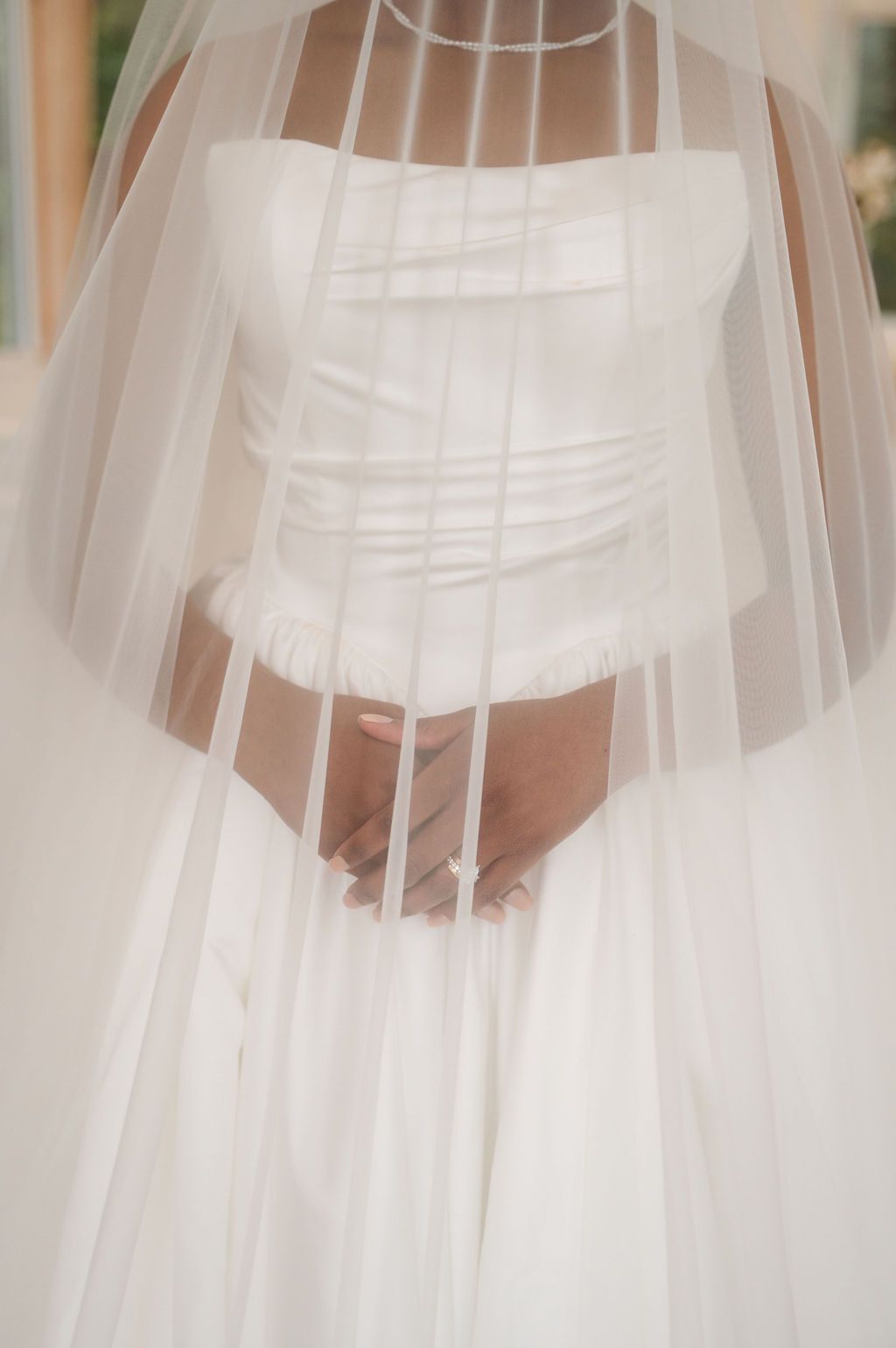 Close-up of bride in strapless white wedding gown with sheer layered veil draped over hands.