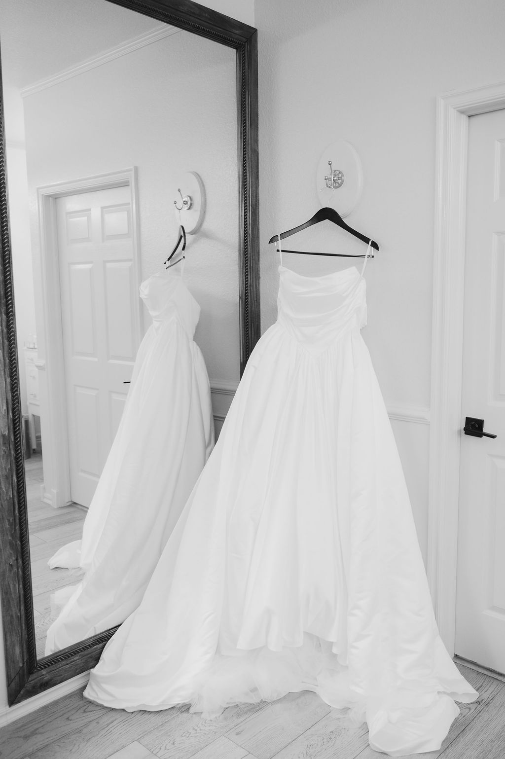 Black and white photo of an elegant strapless wedding ball gown hanging on a hanger beside a large floor mirror.