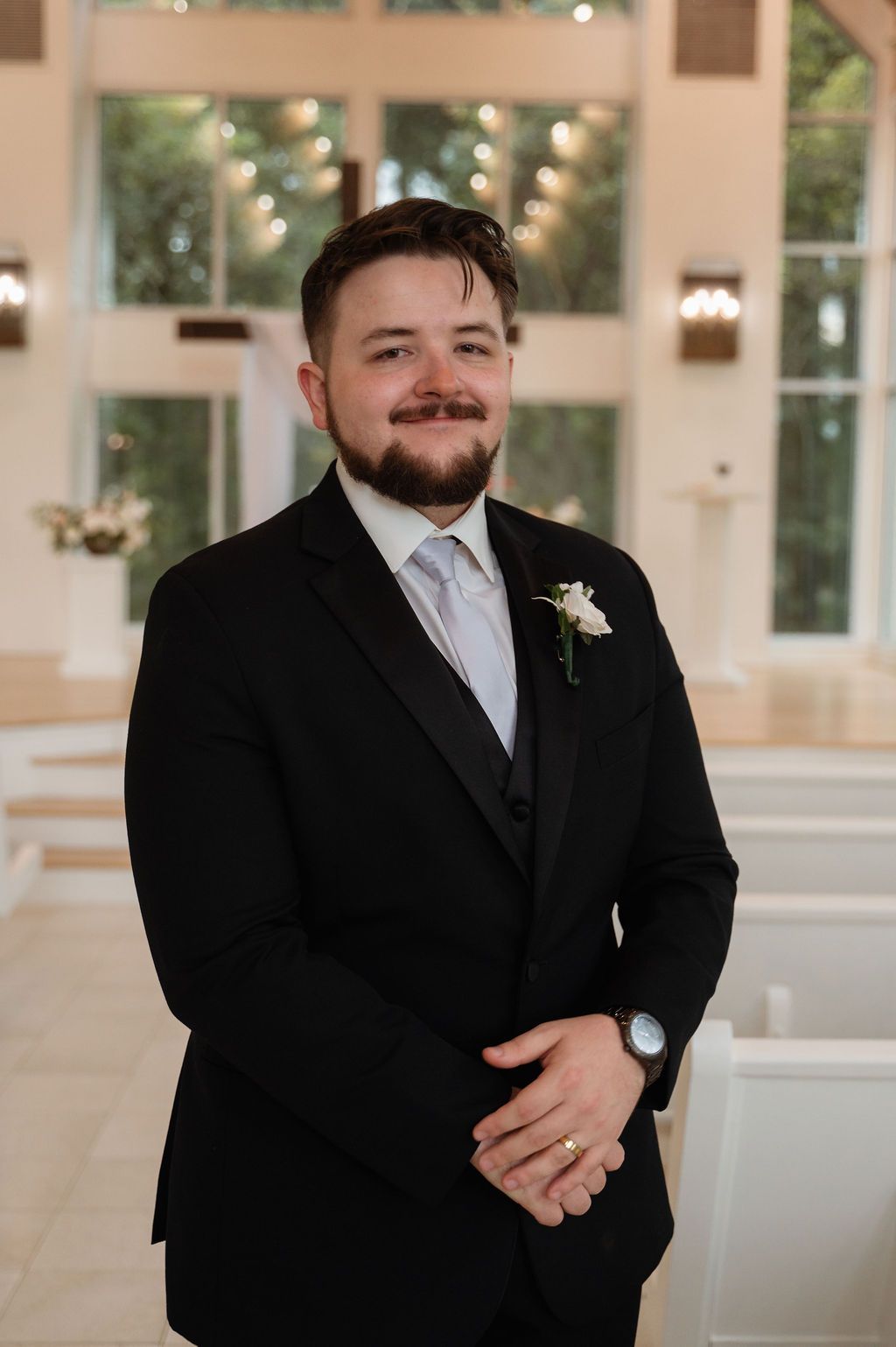Smiling groom in black suit with white boutonniere and gold wedding ring stands in elegant white chapel.