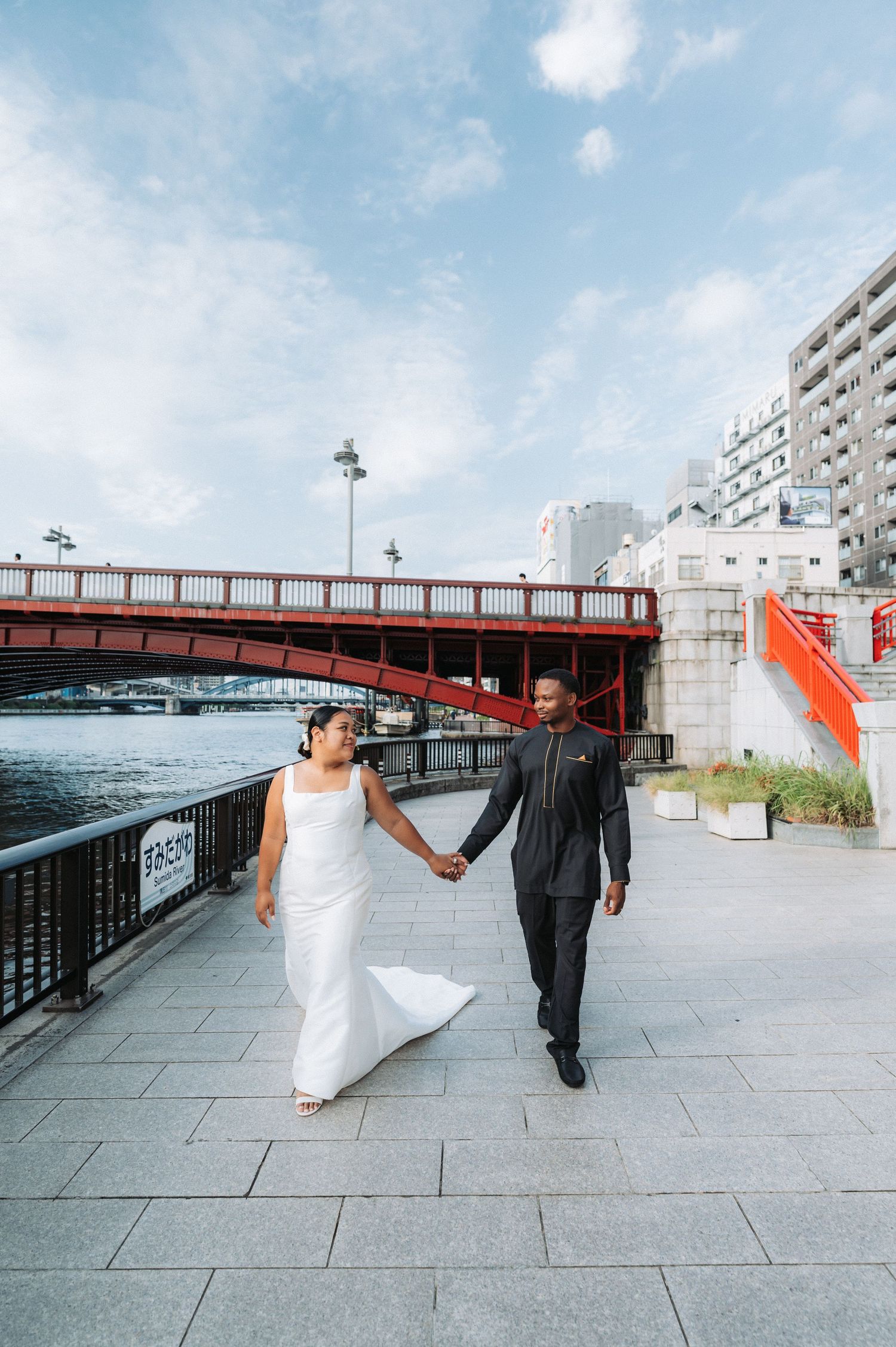 Couple in wedding attire walking along a waterfront pathway with red bridge in background.