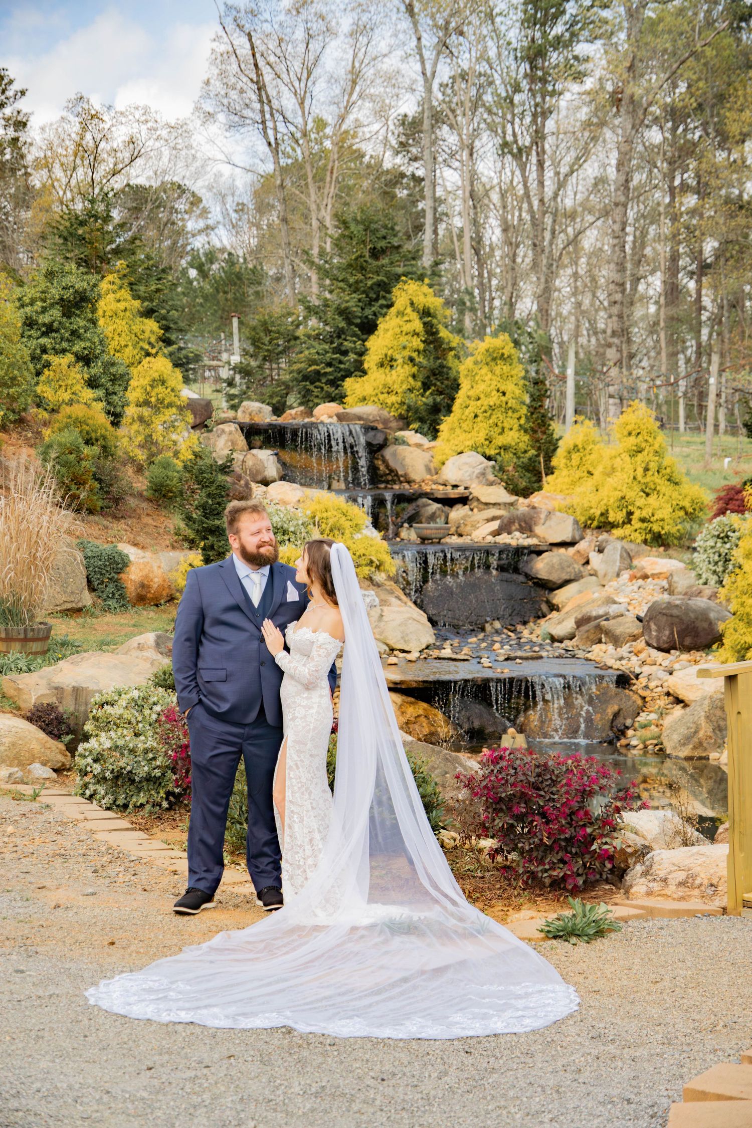 Romantic wedding portrait with flowing cathedral veil among autumn trees and cascading waterfall in garden setting.