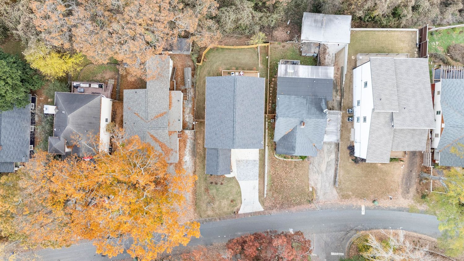 Aerial drone view of residential houses with autumn trees and gray rooftops along a suburban street.