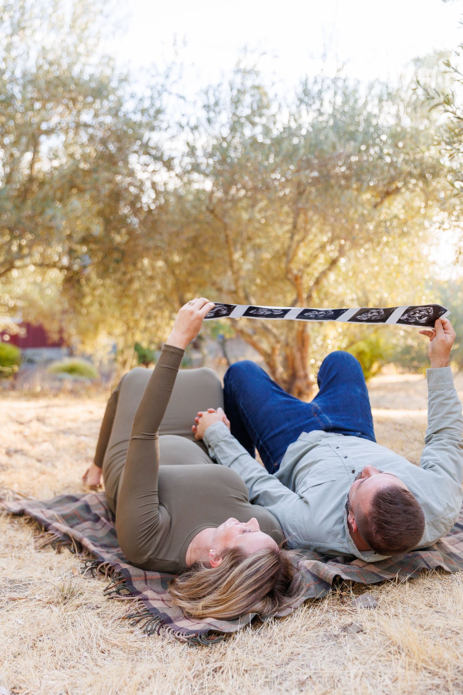 Lily and Travis lie back on a blanket in their Napa olive grove smiling at an image of their sonogram during their maternity session.