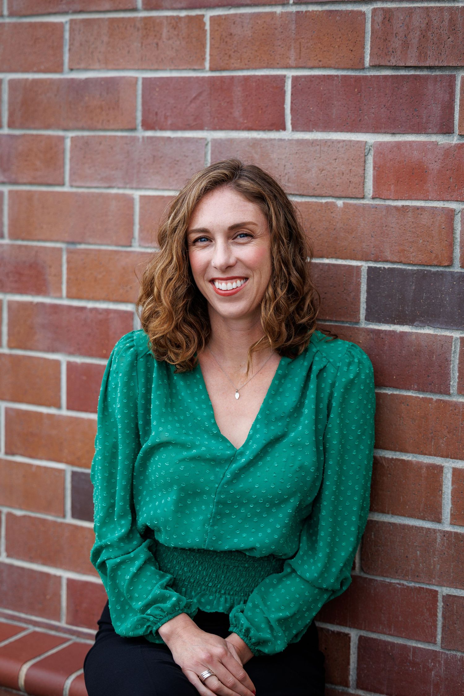 Elly sits against a brick wall, wearing a green blouse and black pants, smiling softly.