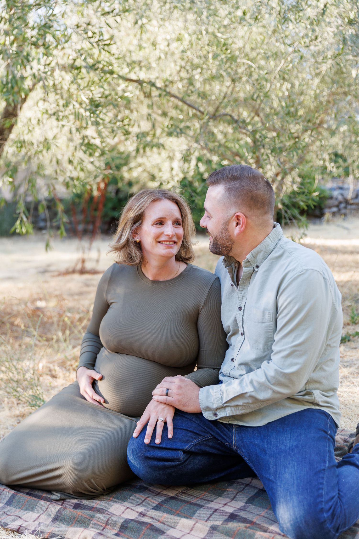 Lily and Travis sit close together in the olive grove, smiling at the camera, with Lily’s baby bump in focus.