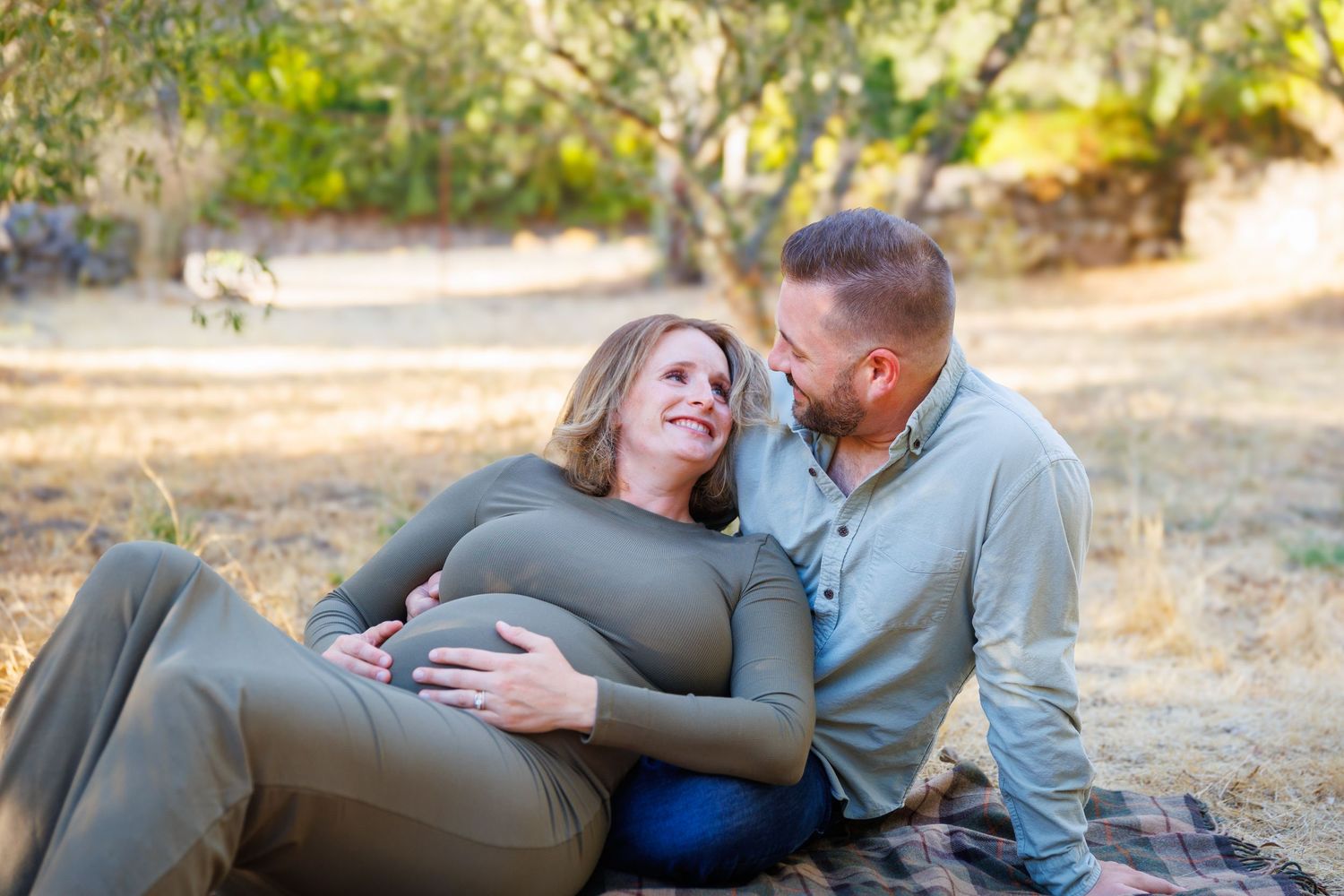 Lily leans back against Travis as they sit on a blanket in the olive grove, laughing together in the warm Napa light.