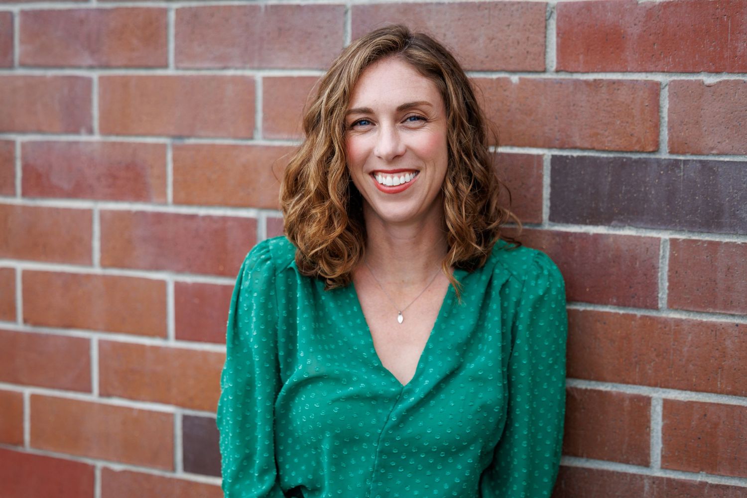 Close-up of Elly smiling in front of a brick wall, wearing a green blouse.