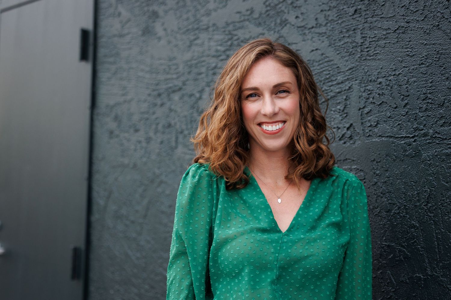 Elly stands against a gray textured wall, smiling warmly while wearing a green blouse.