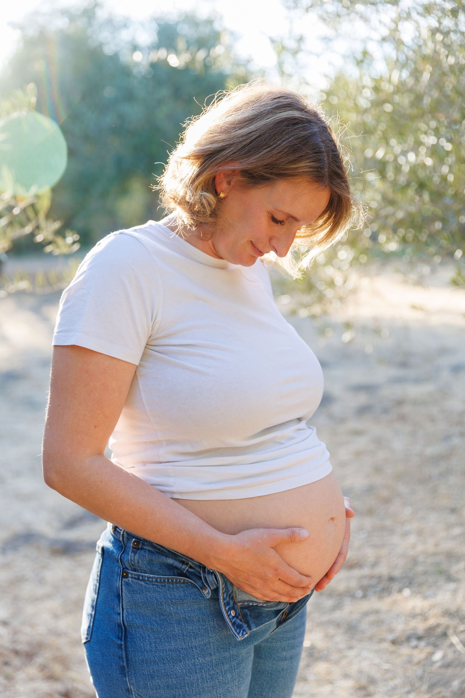 Lily stands in the Napa olive grove, hands resting on her baby bump, smiling softly during her maternity session.