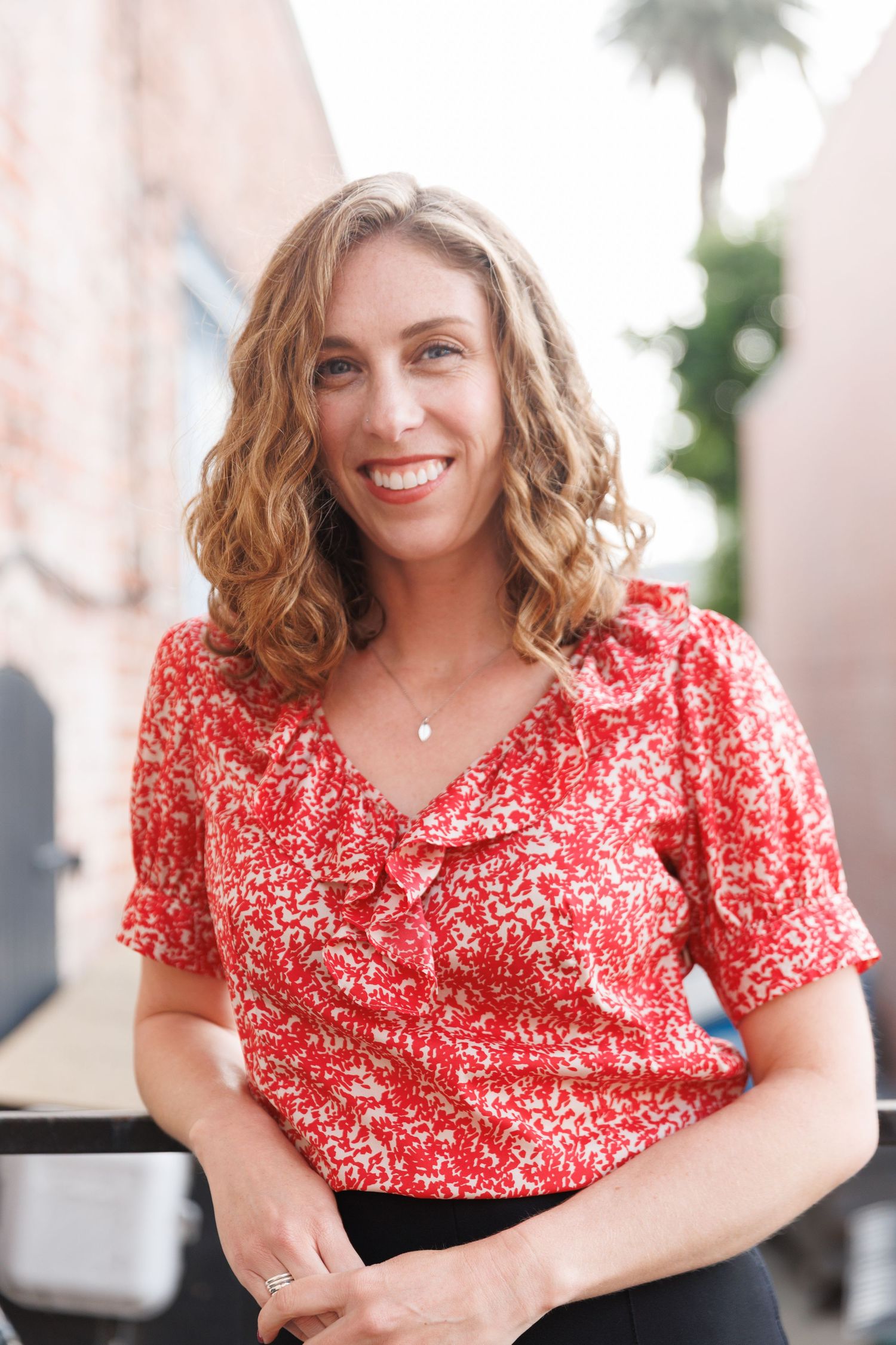 Elly poses outdoors against a light wall, smiling warmly in a red blouse and black pants.