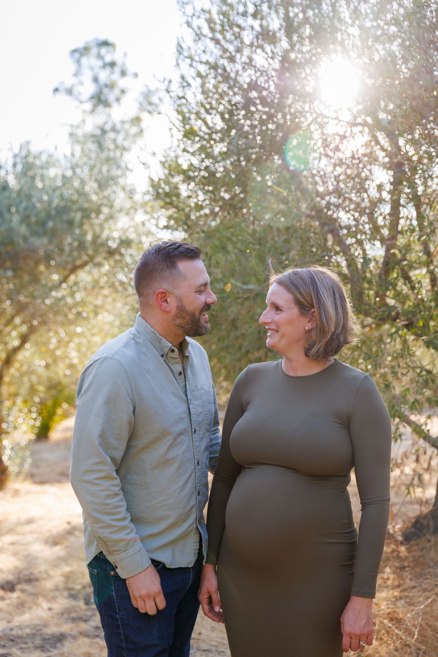 Lily and Travis stand hand in hand in their Napa olive grove at golden hour, smiling and looking at one another.