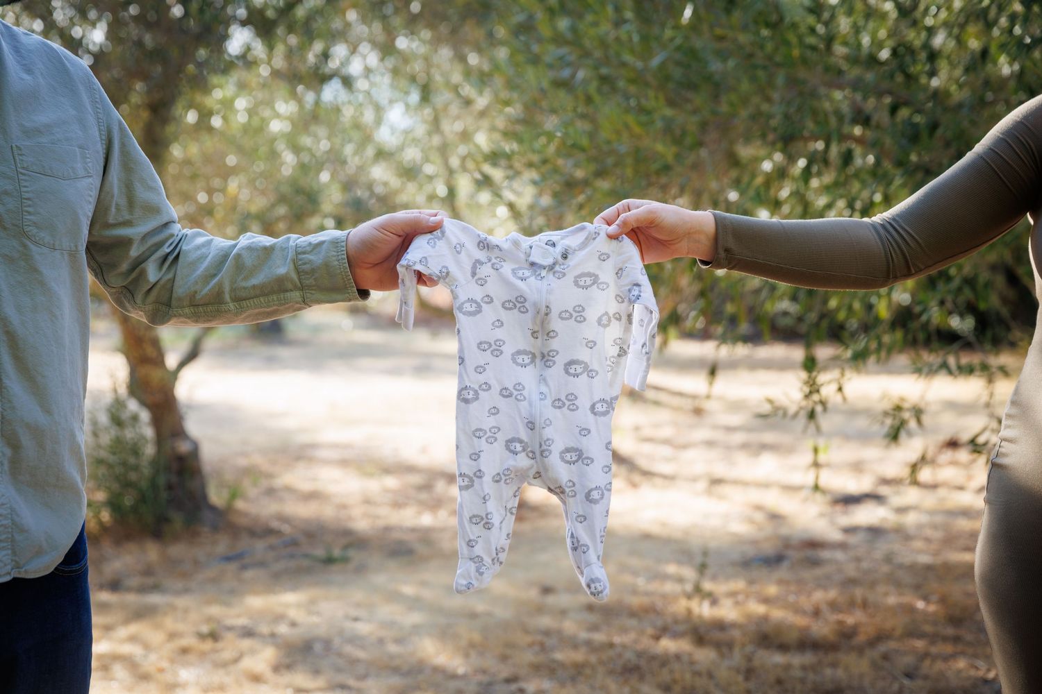 Lily and Travis each hold one side of a tiny patterned baby onesie, stretched between them in the olive grove.