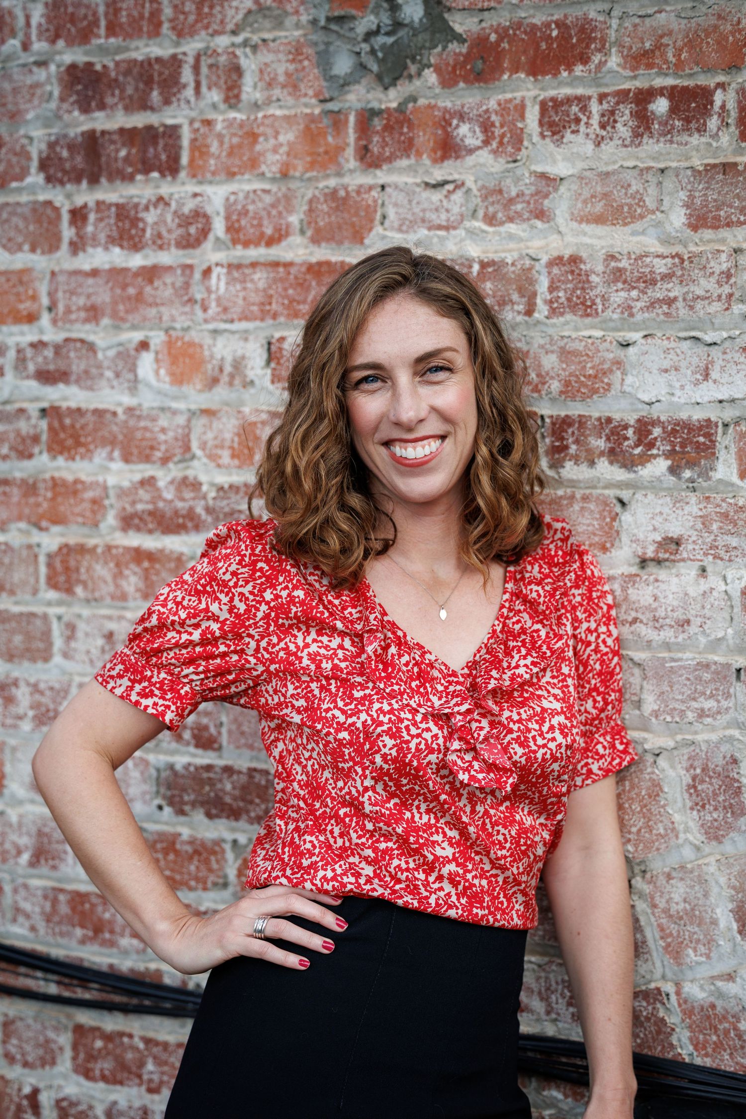 Elly stands in front of a brick wall, hand on hip, wearing a red blouse and smiling at the camera.