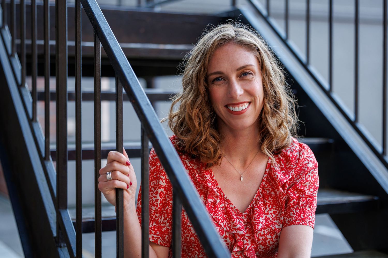 Elly smiles while sitting on outdoor stairs, wearing a red patterned blouse and looking confident.