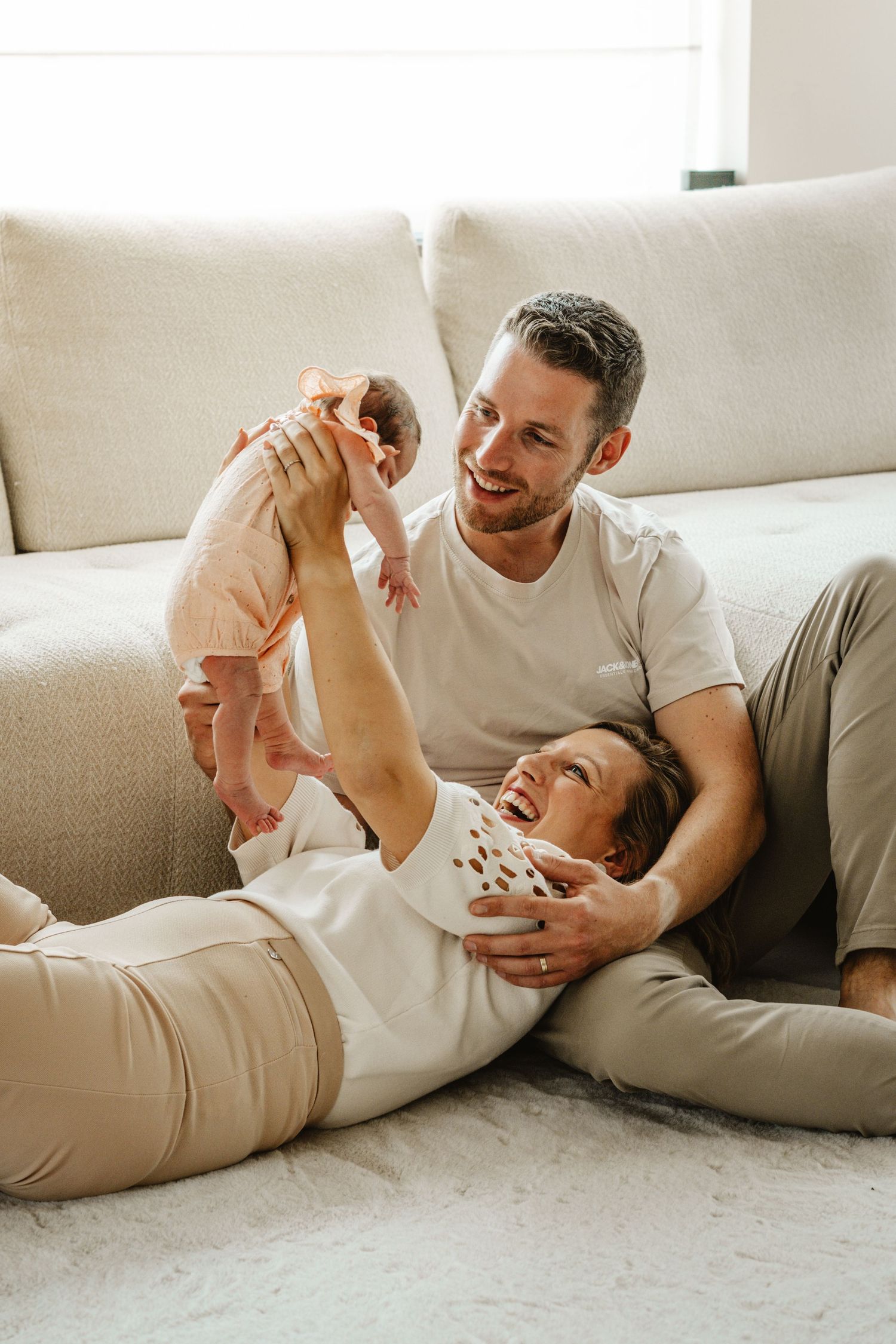 Parents playing with infant while lying on white couch in natural light.
