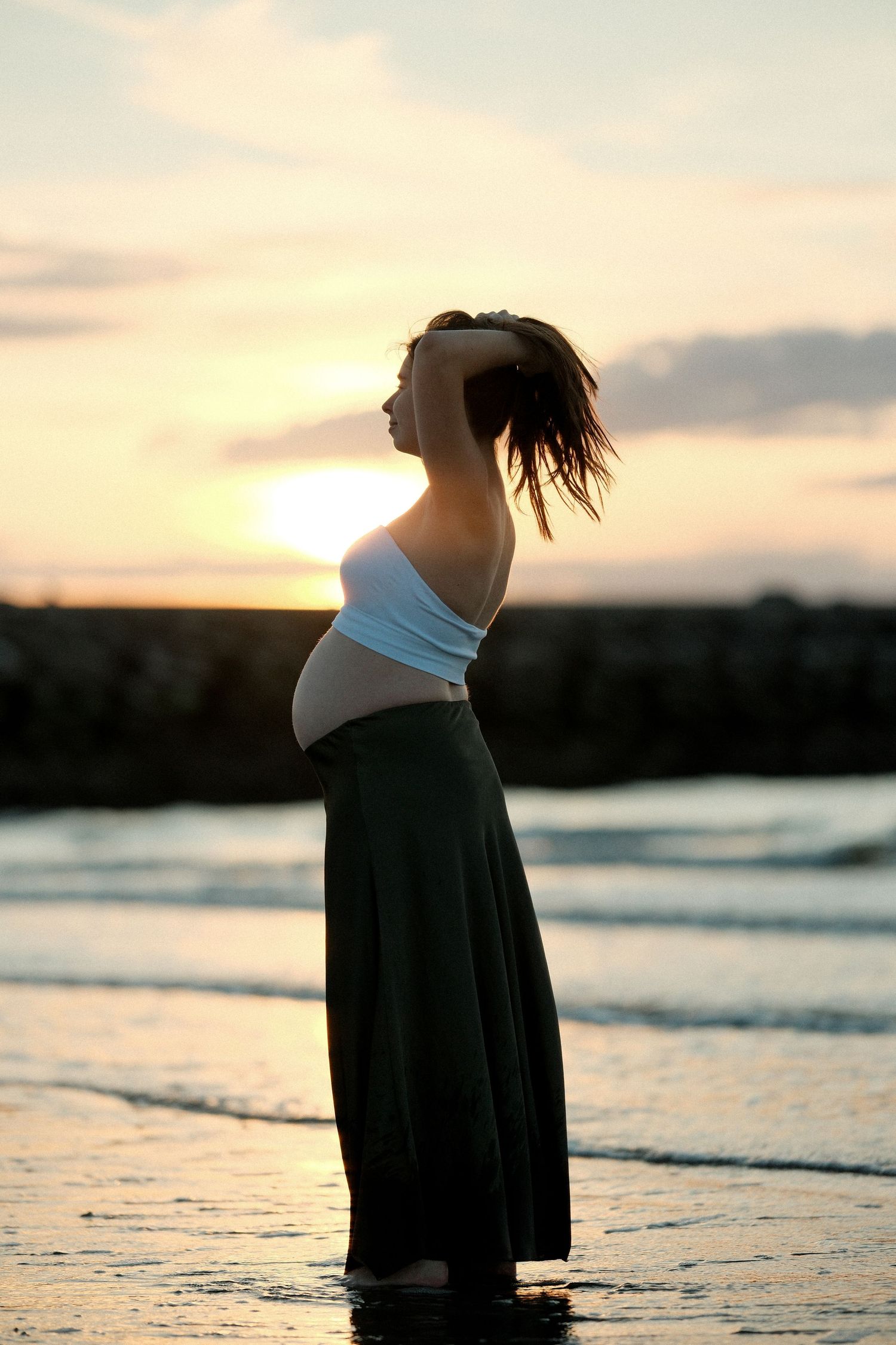 Pregnant woman standing barefoot on a beach at sunset, wearing a white top and dark skirt, hair blowing in the wind.