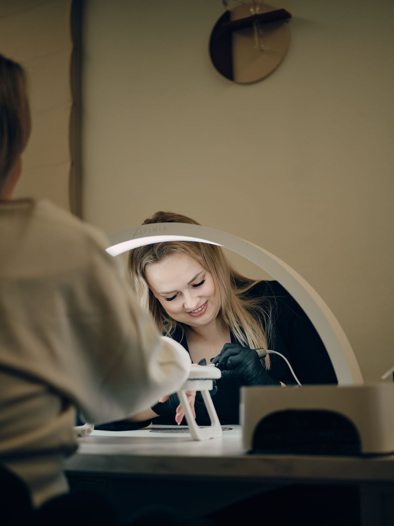 A smiling nail technician in black gloves works on a client's nails under a magnifying lamp in a modern salon.