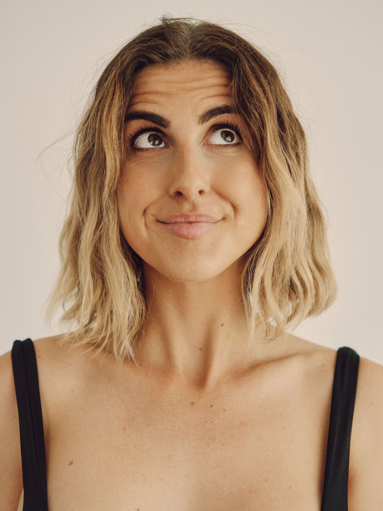 Young woman with blonde bob hair making a playful expression, looking up against a light background.