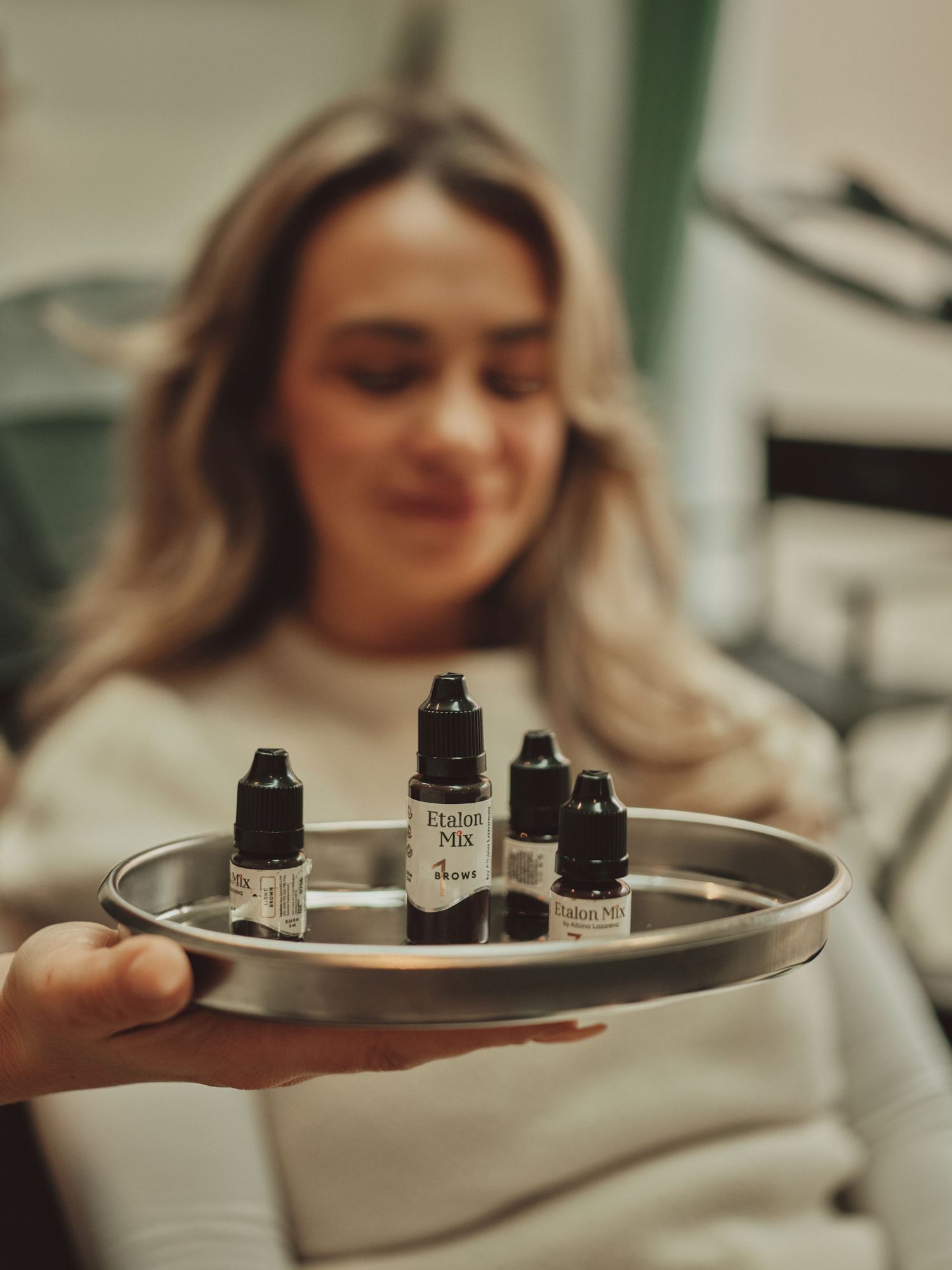 Woman smiling behind a silver tray holding four small Elixir Mix serum bottles in a bright salon setting.