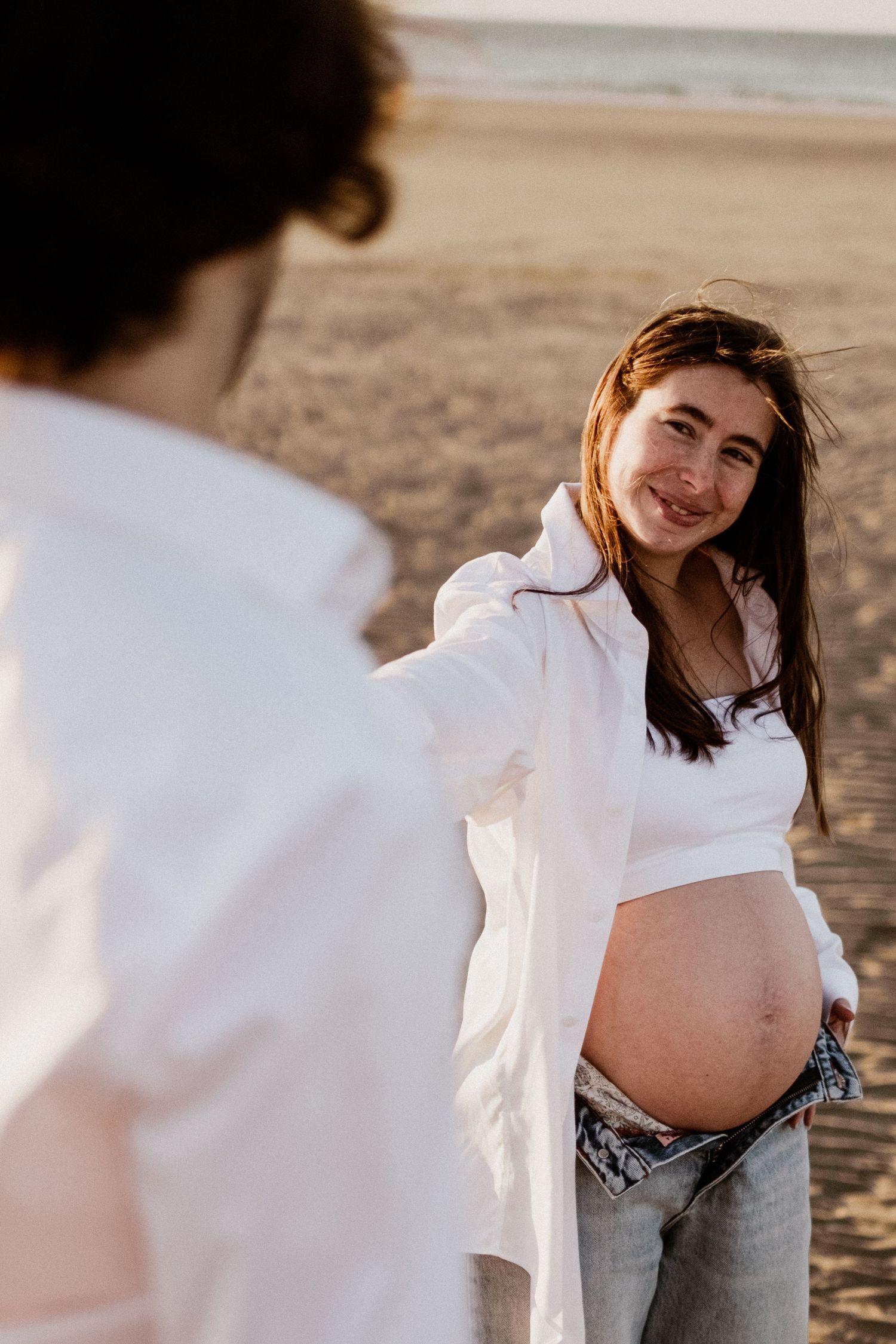 Candid maternity beach photos at sunset wearing white button-down shirt.