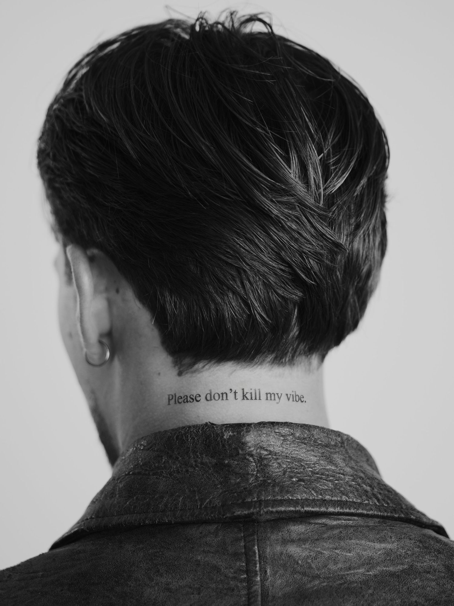 Black and white photo of person with neck tattoo reading please don't kill my vibe, wearing leather jacket.
