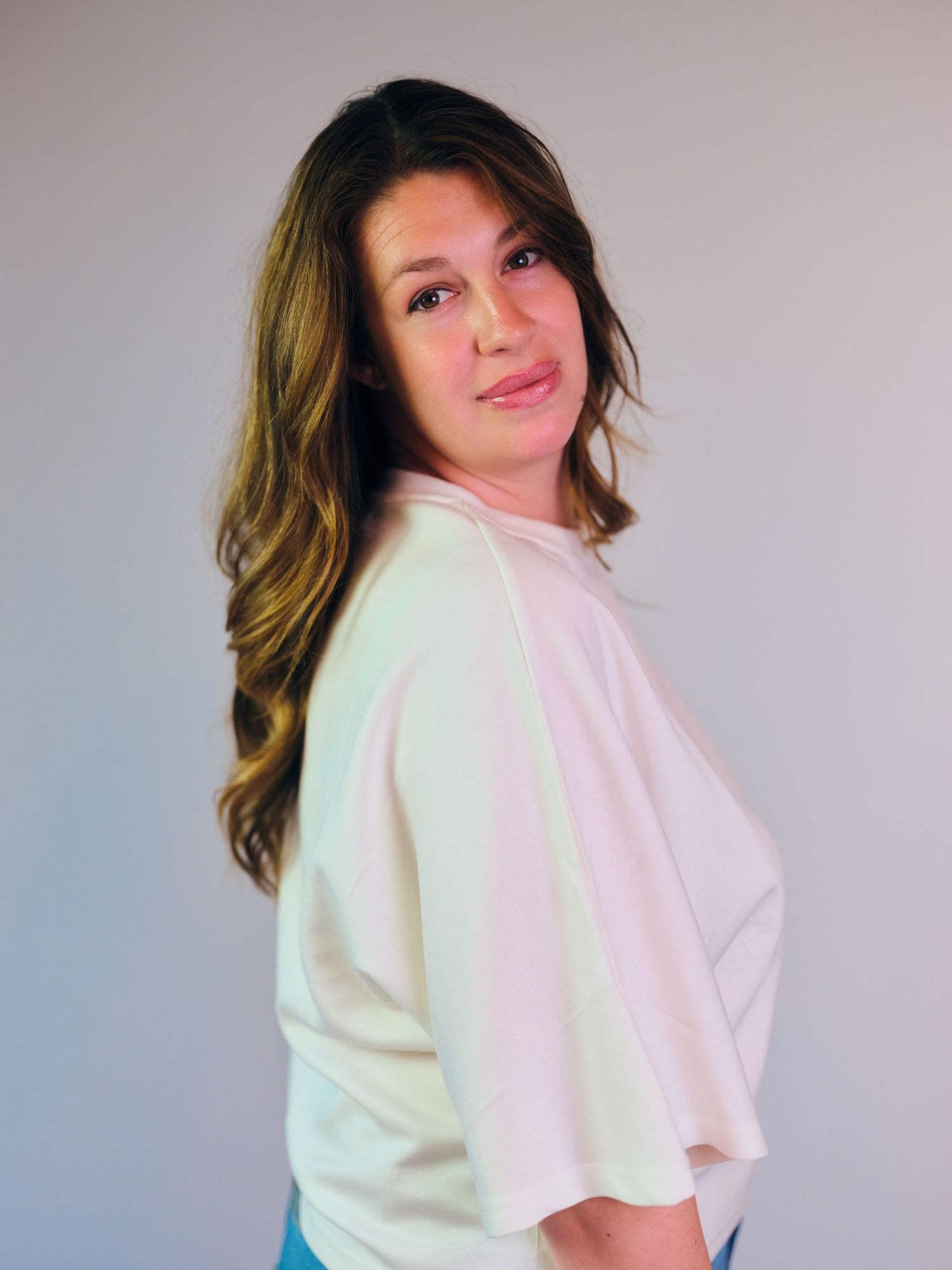 Woman with long brown hair poses sideways in a white top against a gray background.