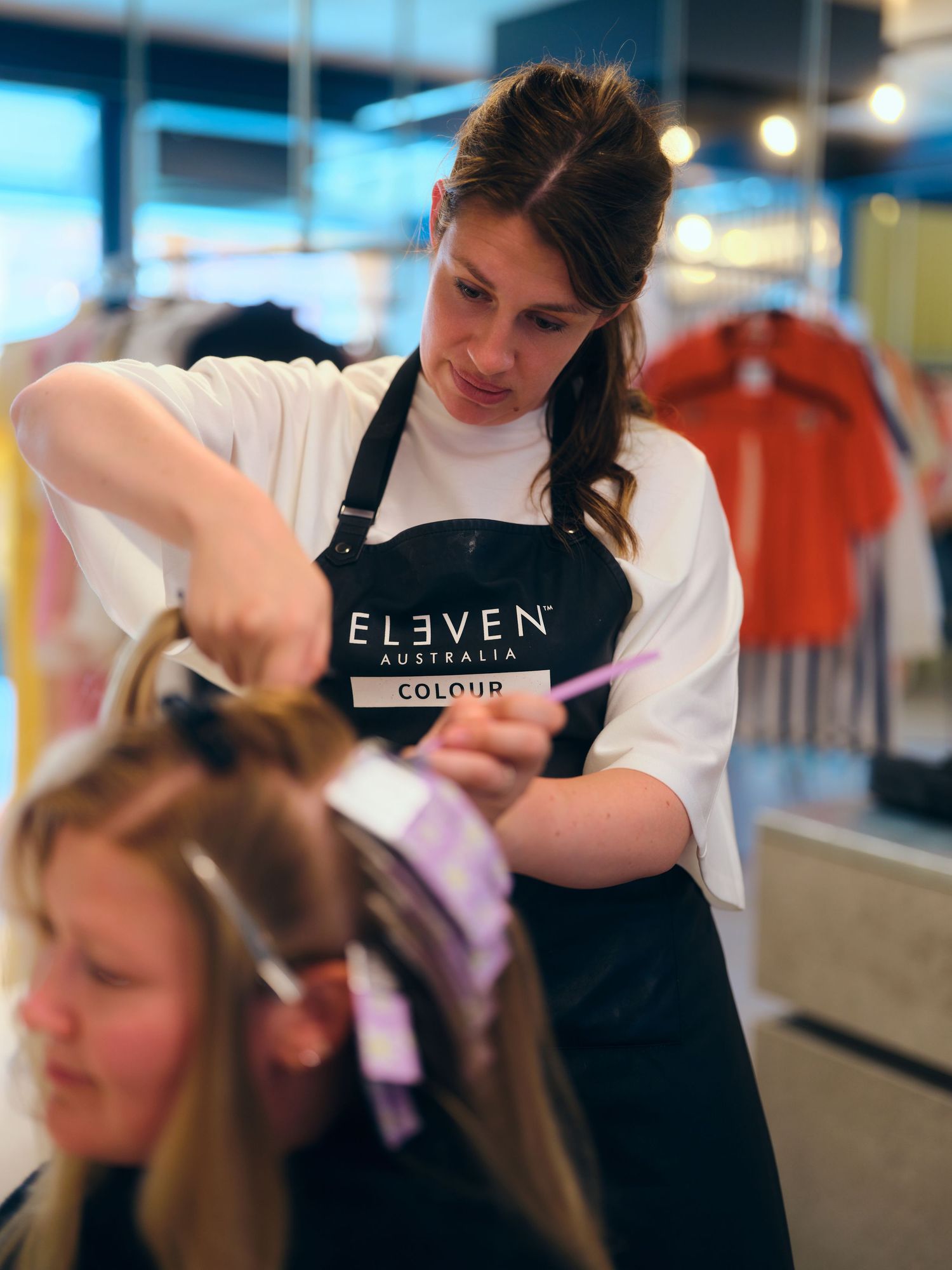 Hairstylist wearing Eleven Australia Colour apron applies hair dye to a blonde client in a modern salon.