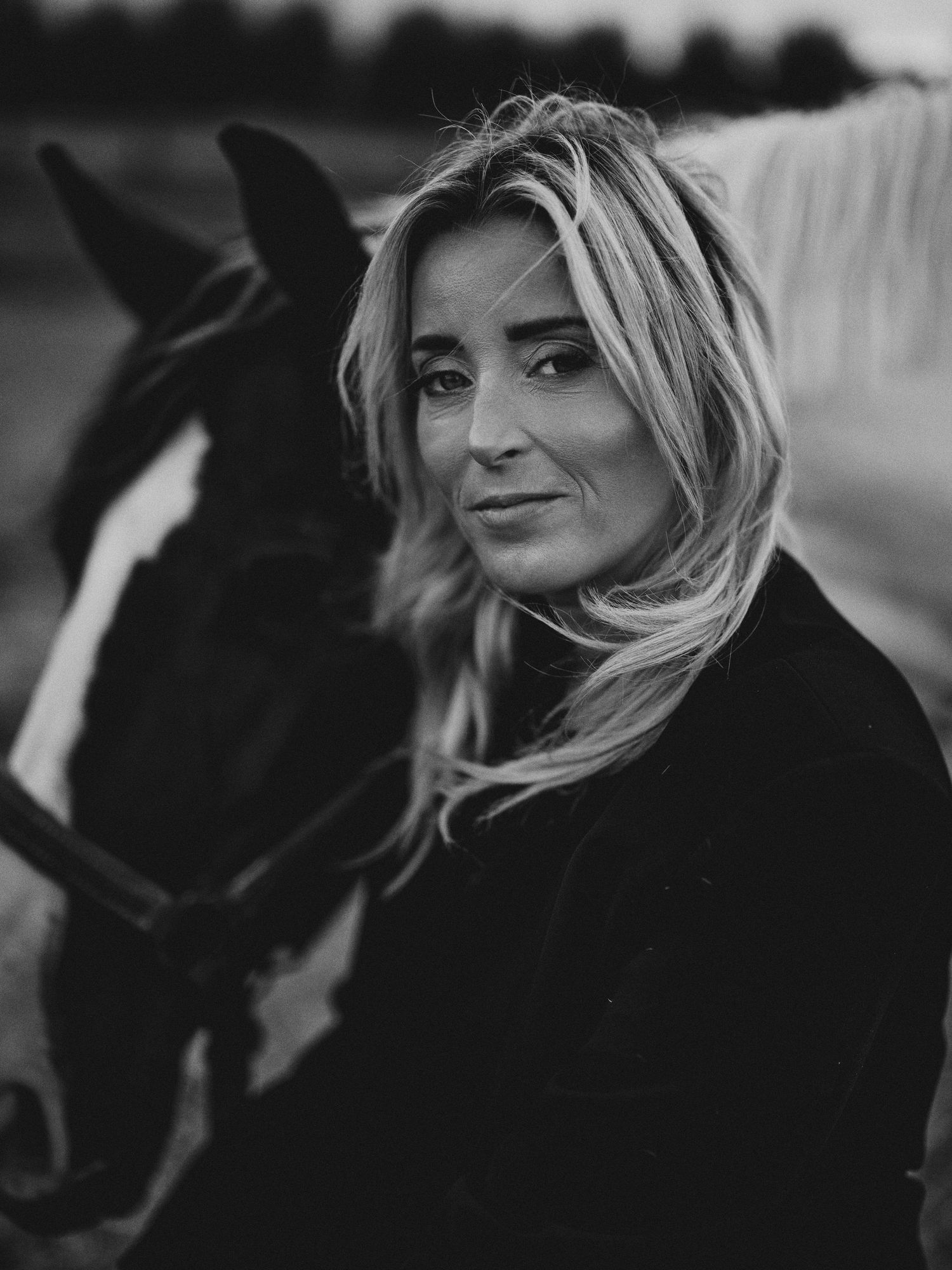 Black and white portrait of a blonde woman posing with a horse outdoors.