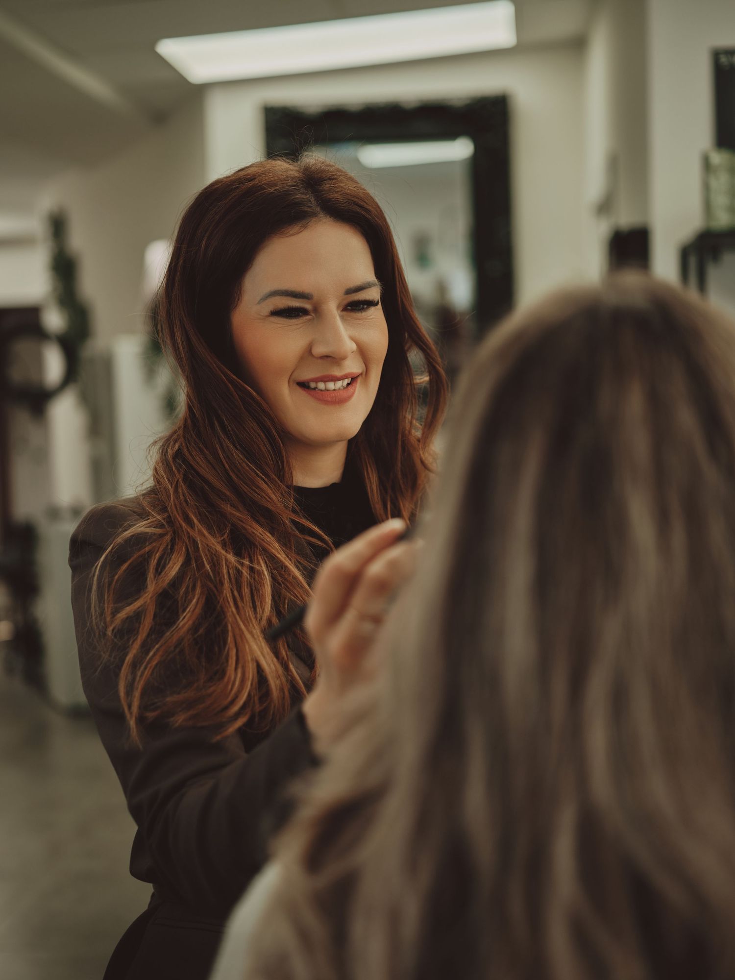A smiling brunette hairstylist in a black top consulting with a client in a modern hair salon.