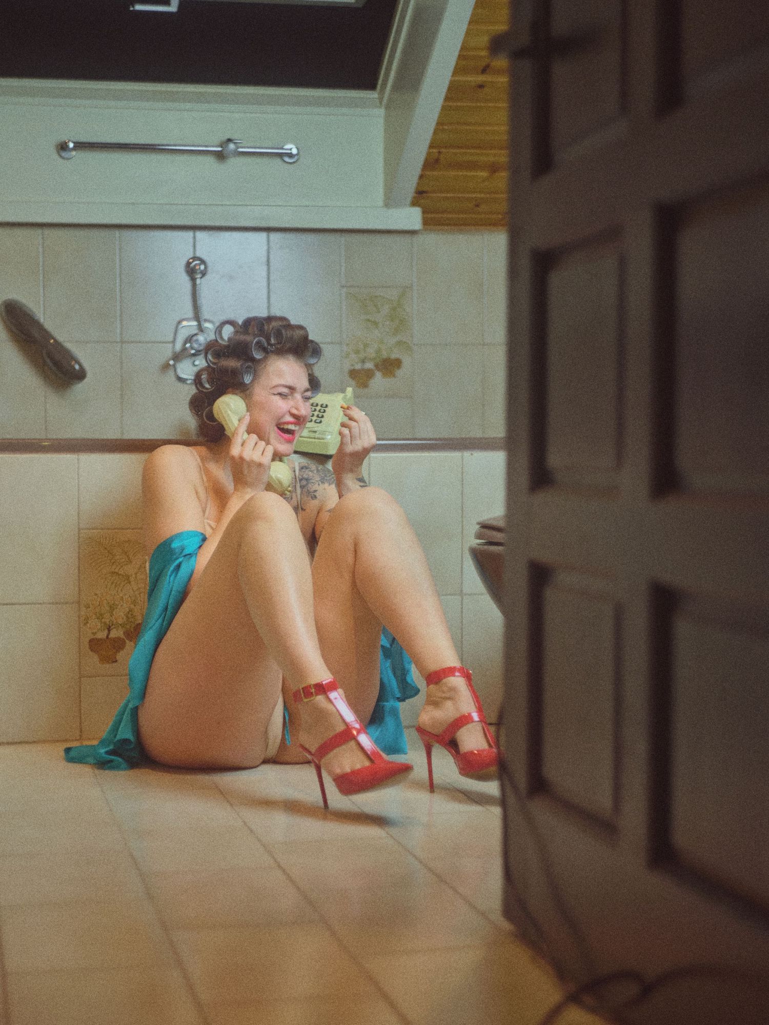 Woman in red heels sitting on bathroom floor laughing while talking on a vintage green telephone.