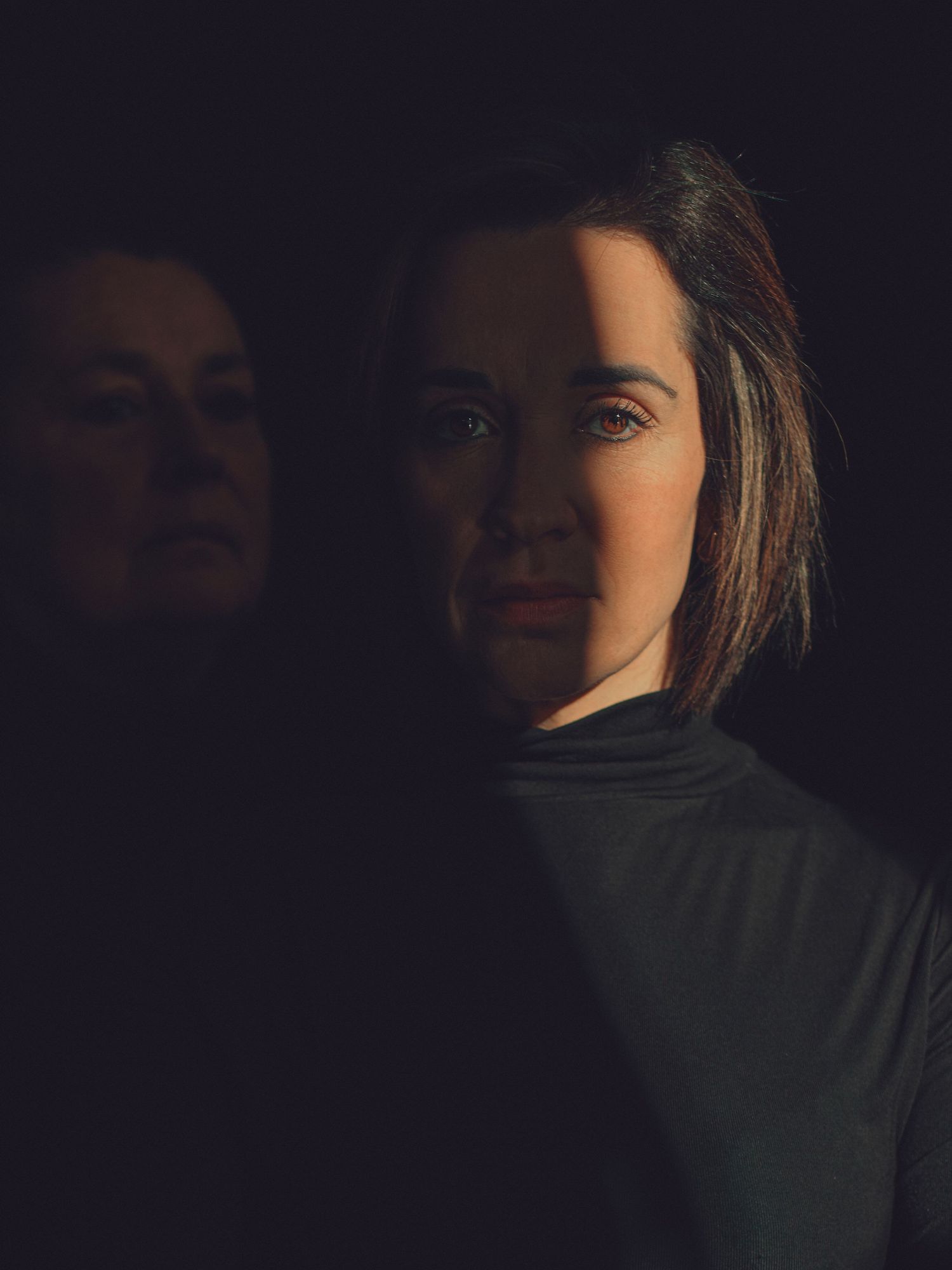 A woman in a dark turtleneck with dramatic lighting, her reflection visible in a mirror behind her.
