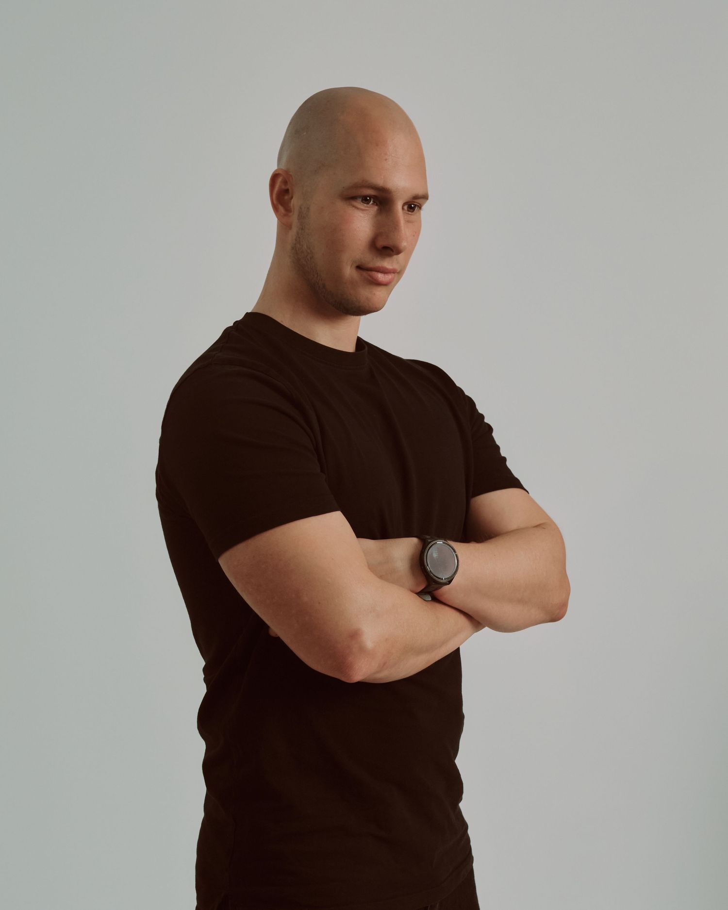 Person in brown t-shirt standing with arms crossed against light background wearing watch.