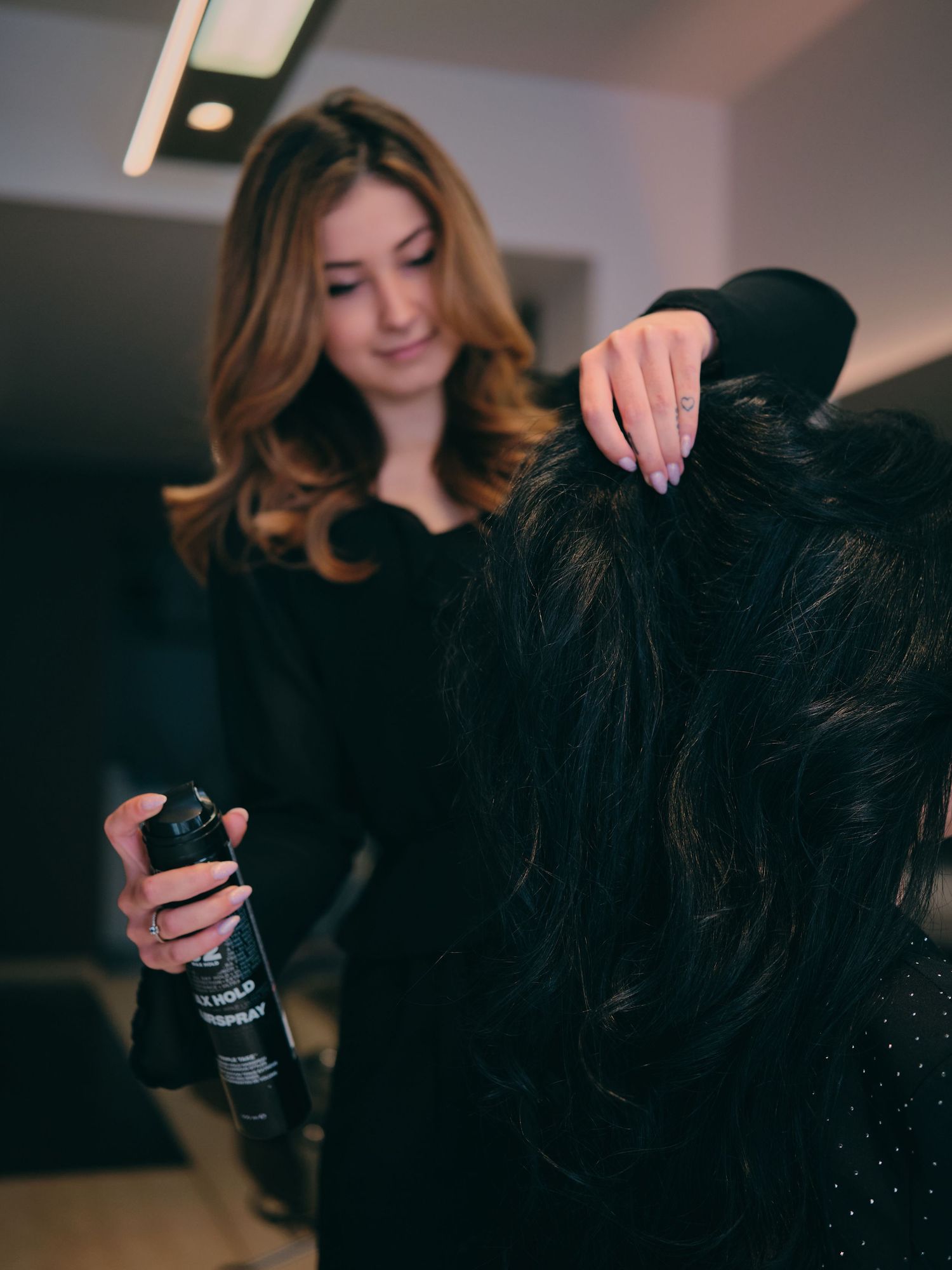 A female hairstylist applies hairspray to a client with dark hair in a modern salon setting.