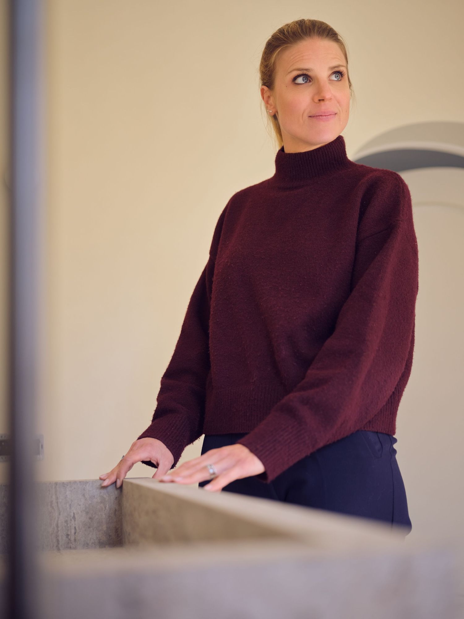 Woman in burgundy turtleneck sweater standing thoughtfully, looking upward in a minimalist interior setting.