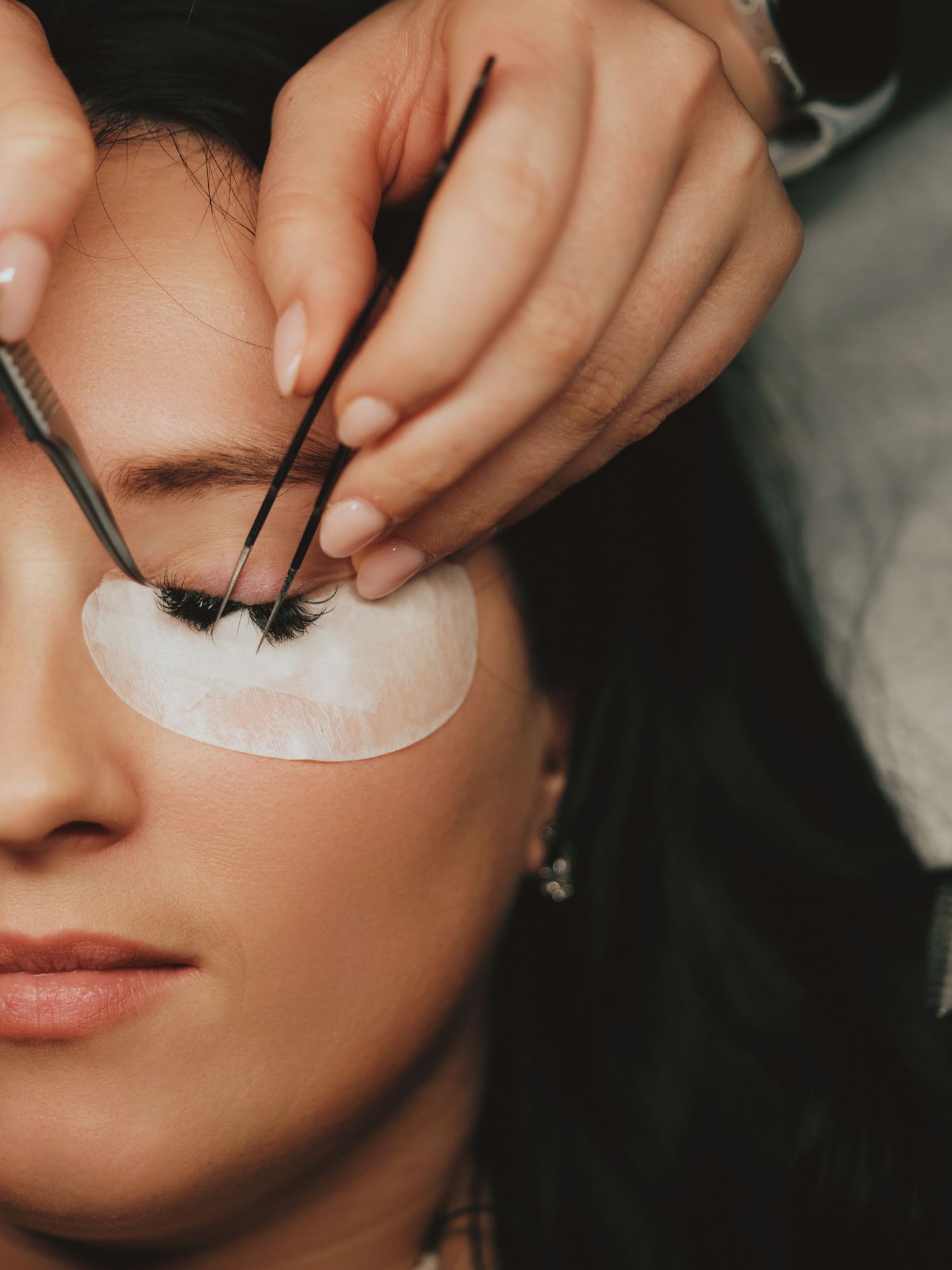 Esthetician applying eyelash extensions to a woman with an eye pad, using tweezers for precision placement.