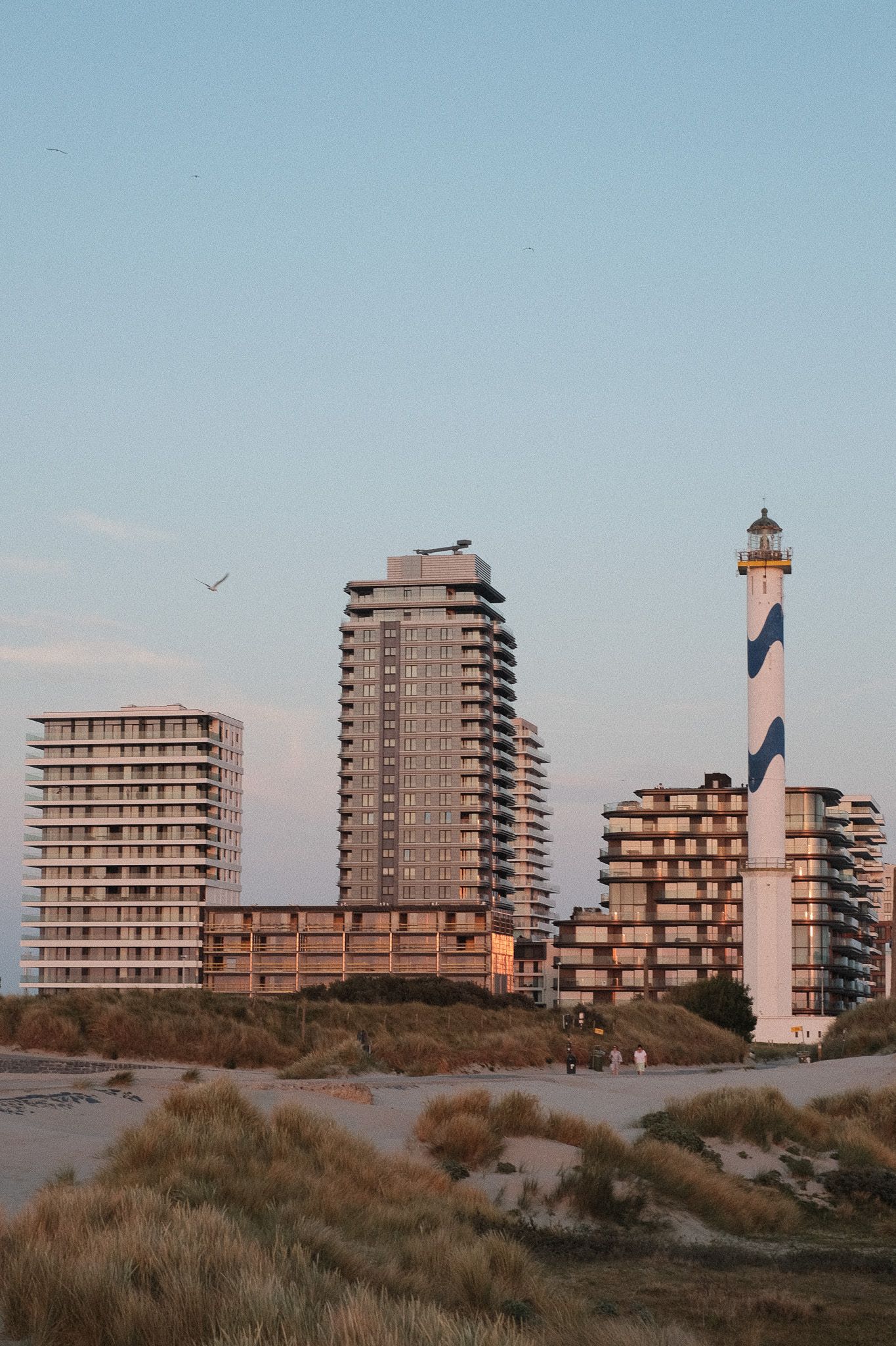 Modern apartment buildings and a lighthouse along coastal dunes at sunset.