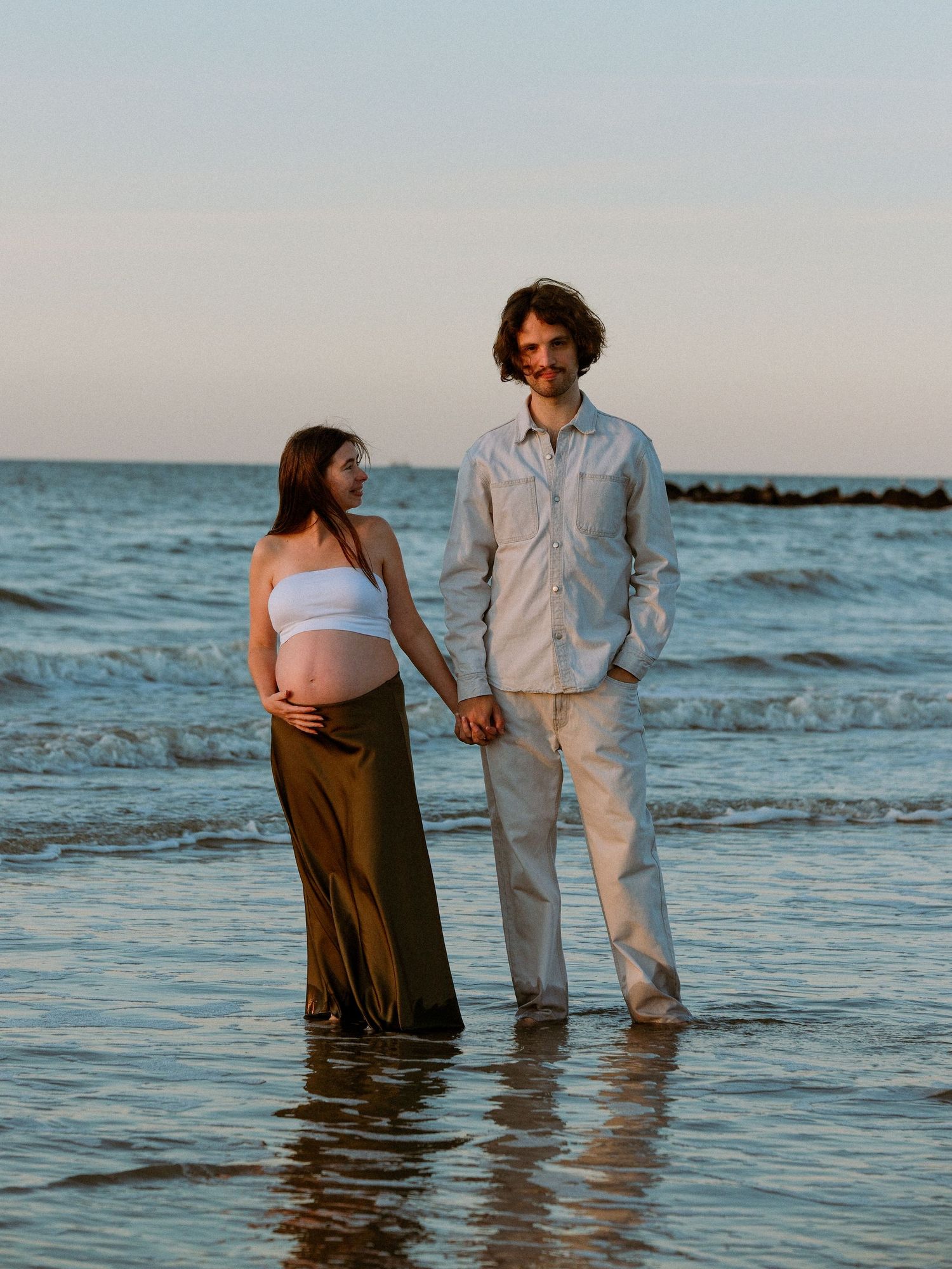 Couple standing together in ocean waves during sunset maternity shoot.