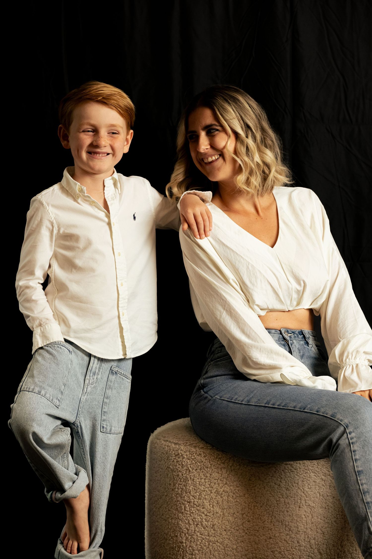 Two people in white shirts and jeans pose together on beige ottoman against dark backdrop.