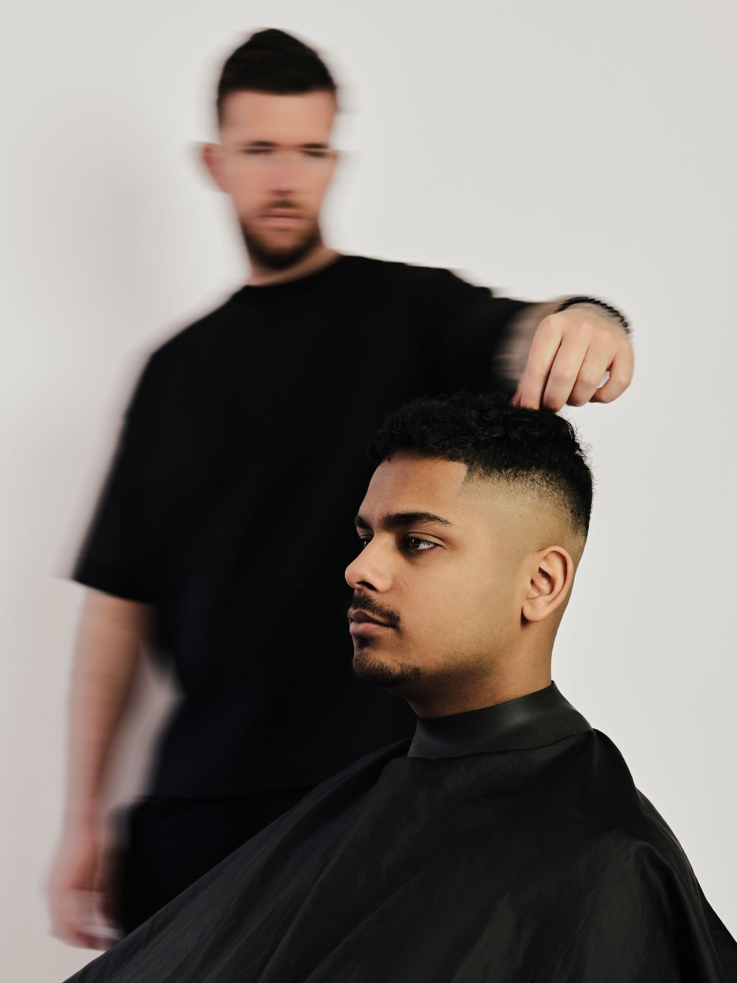 Barber in black t-shirt giving a fade haircut to a young man wearing a black cape in a modern barbershop.
