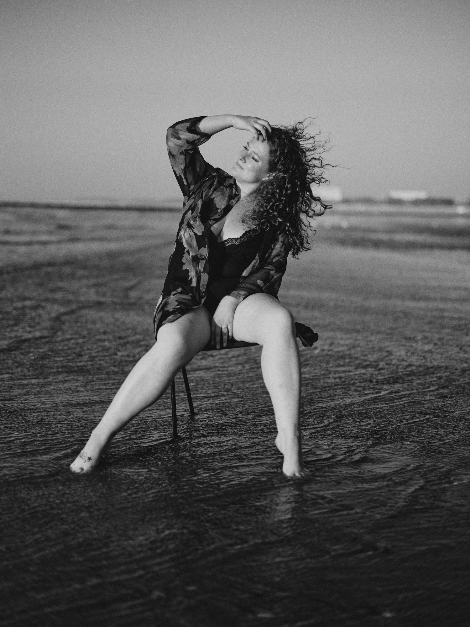 Black and white photo of a curly-haired woman posing on a chair on a wet beach, hair blowing in the wind.