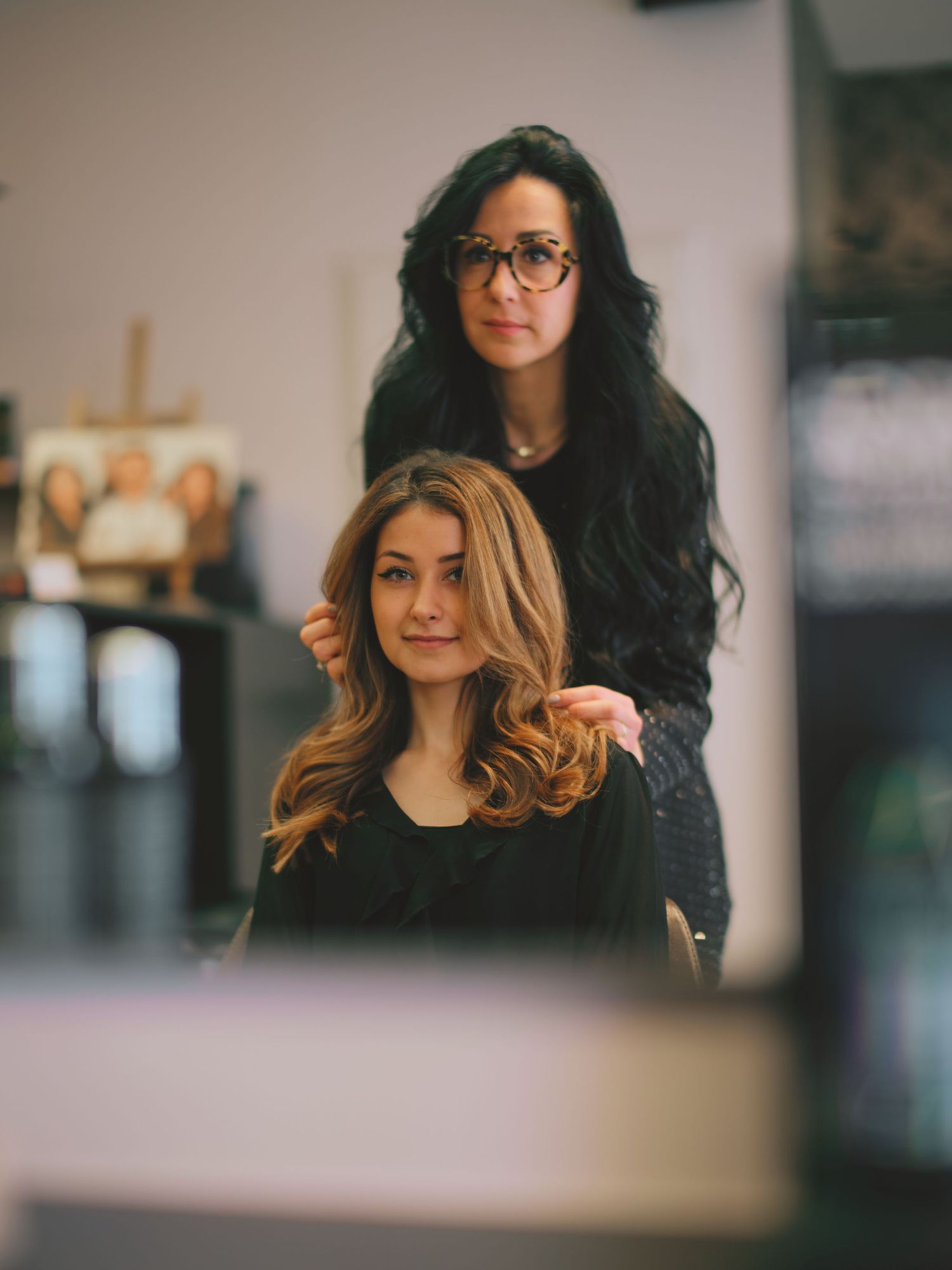 A hairstylist with long black hair and glasses styles a blonde client's wavy hair in a modern salon.