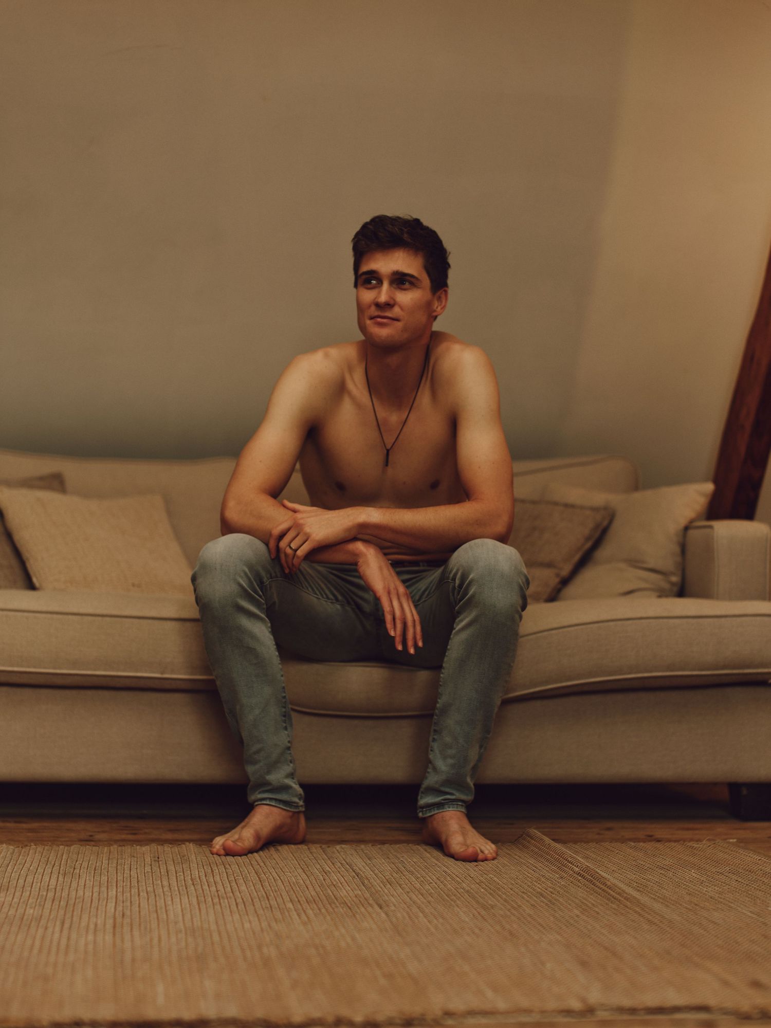 A shirtless person sits casually on a beige couch wearing jeans and a necklace in a warm-toned indoor setting.