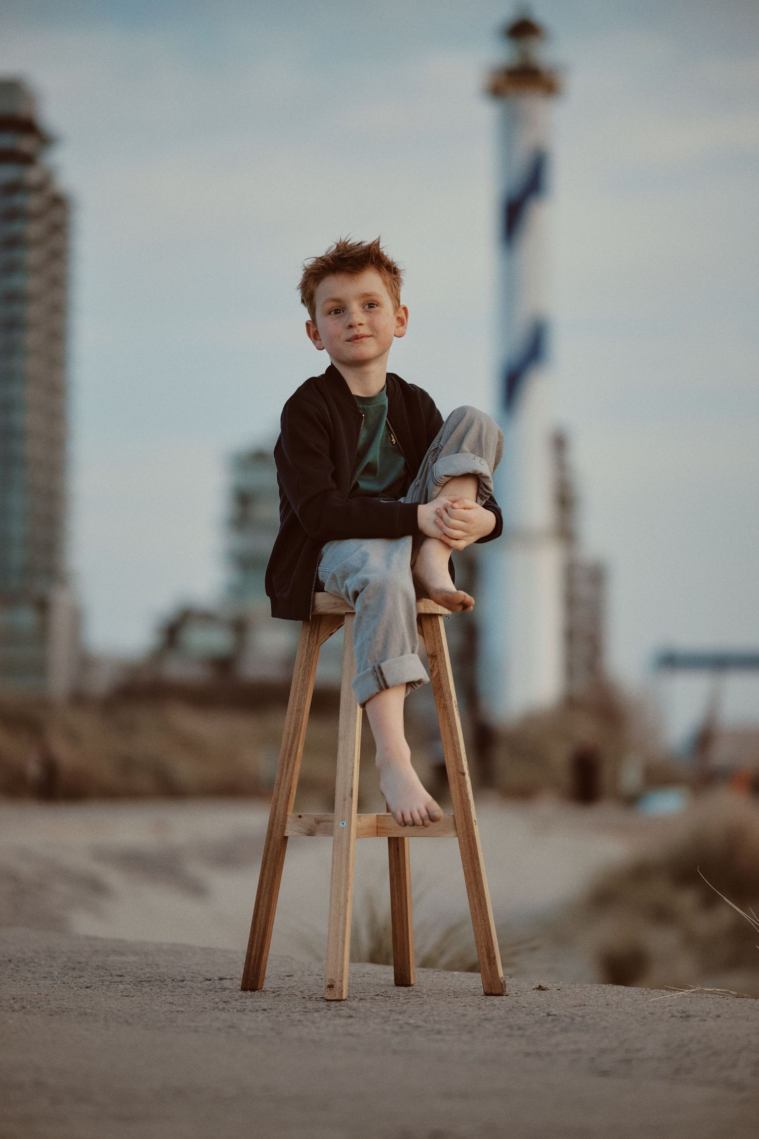 A barefoot boy sits on a wooden stool on a sandy beach with a lighthouse in the background.