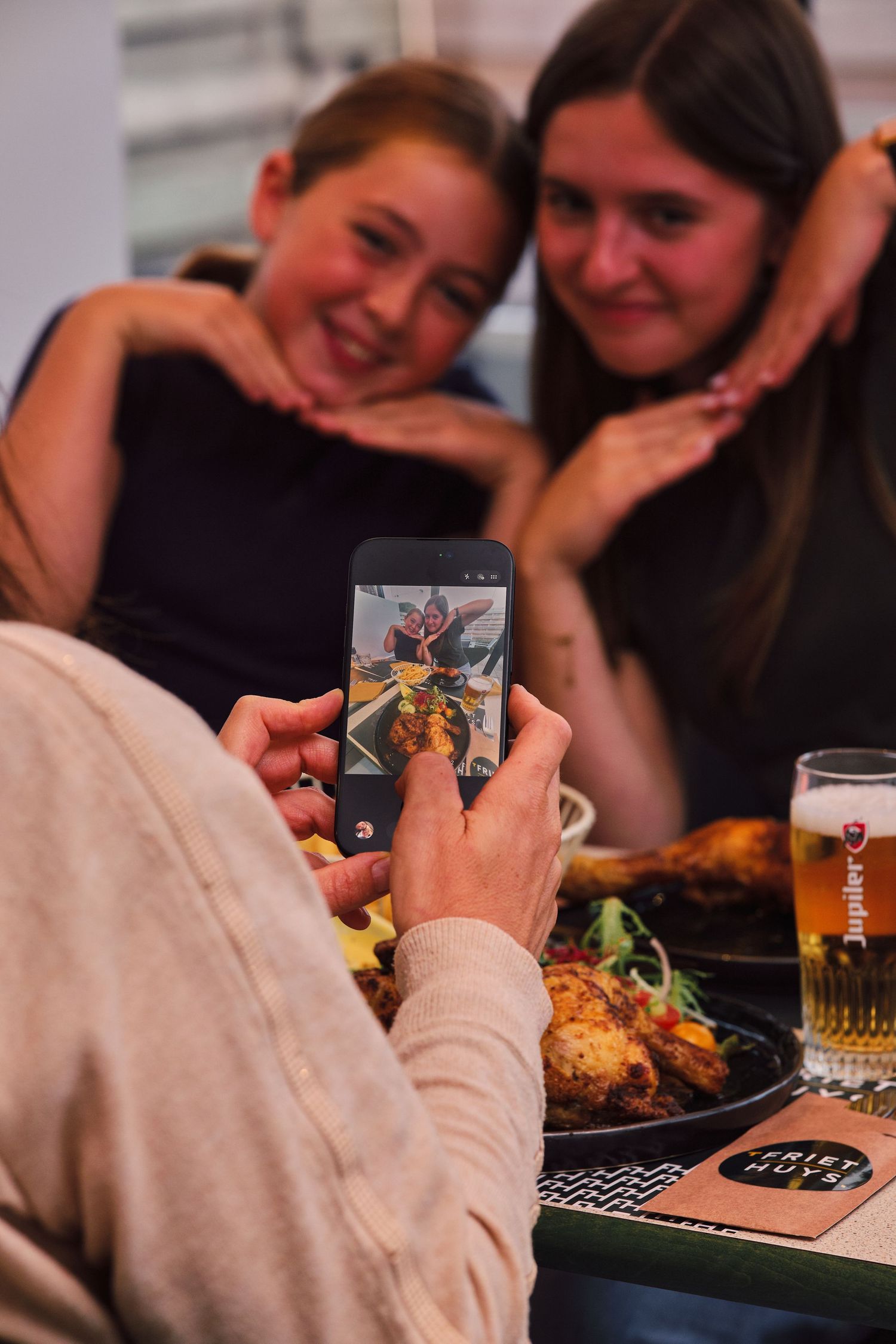 Two women posing for a smartphone photo at a restaurant table with food and a glass of beer nearby.