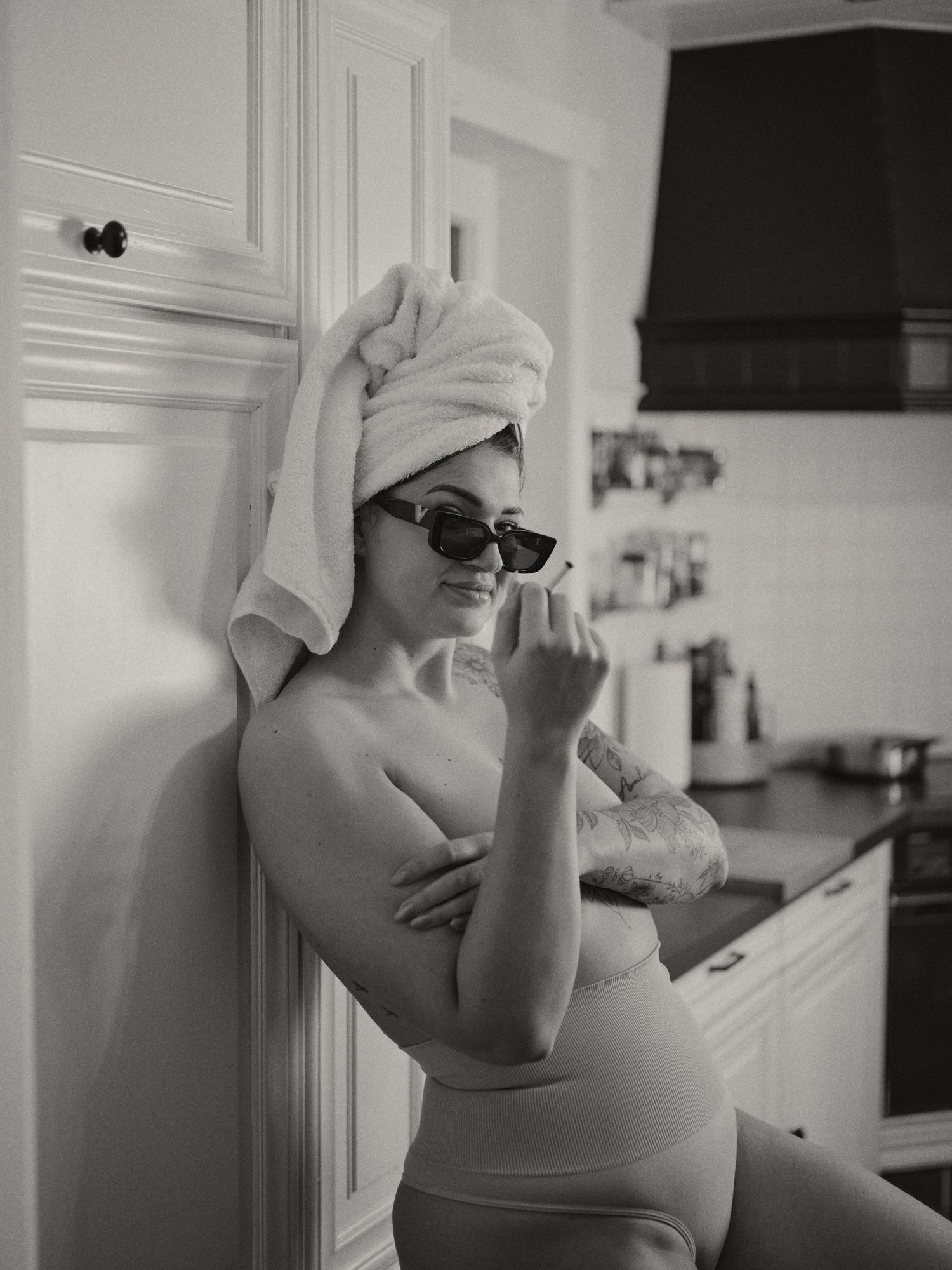 Black and white photo of a woman in a towel wrap and sunglasses leaning against kitchen cabinets.