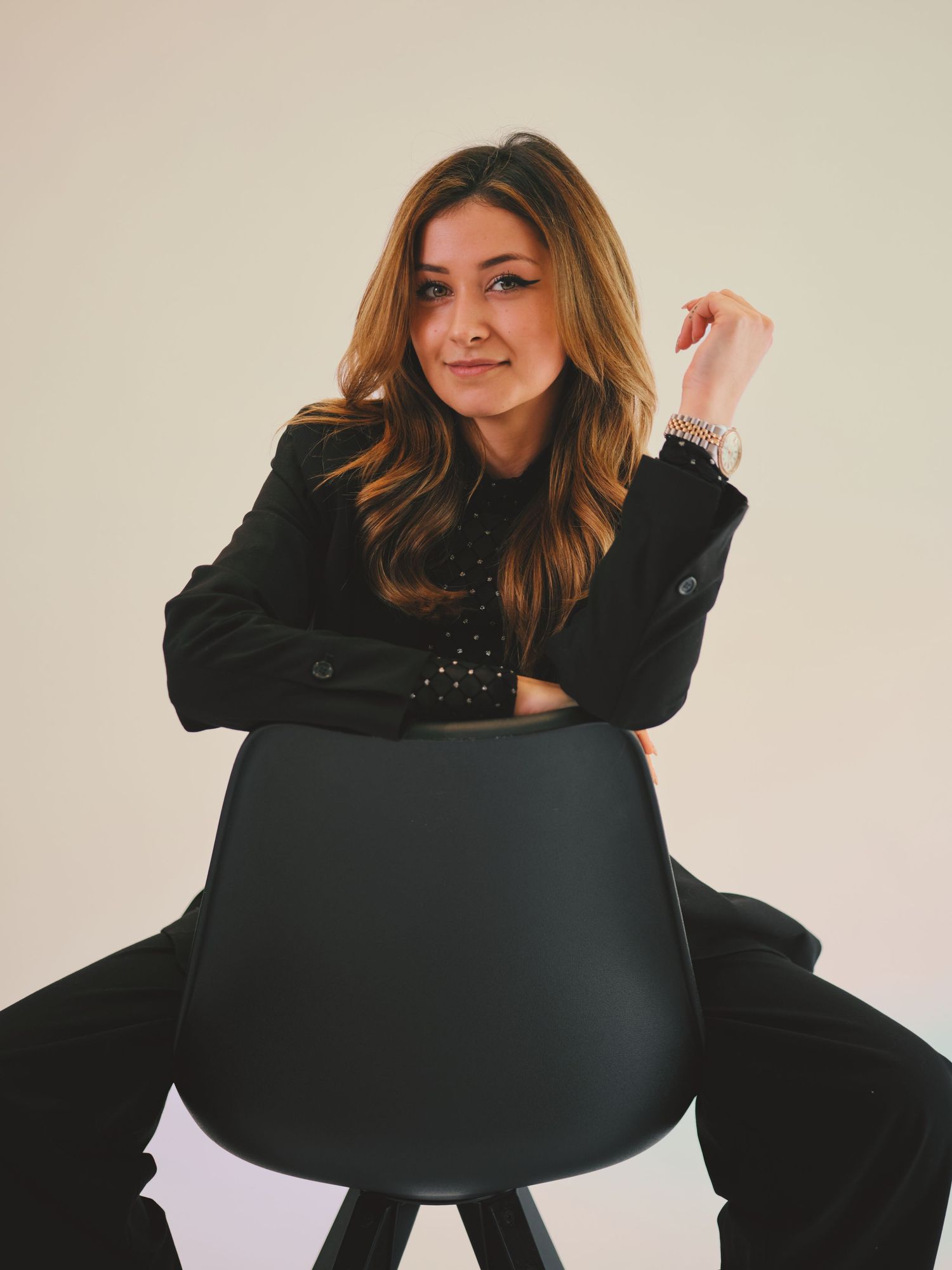 Woman in black blazer posing confidently with arms crossed on a modern black chair against a white background.