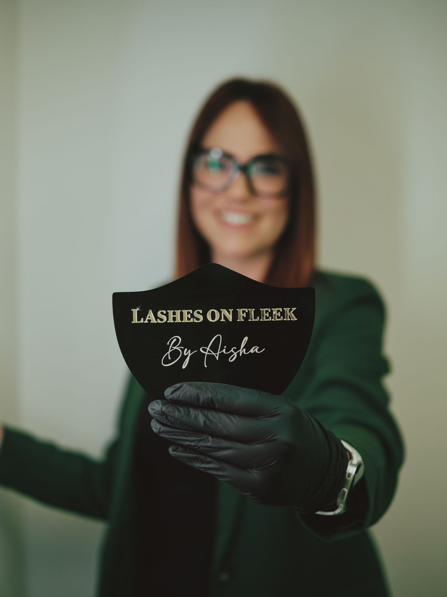 Woman in green blazer holding a black fan-shaped sign reading Lashes On Fleek By Aisha while wearing black gloves.