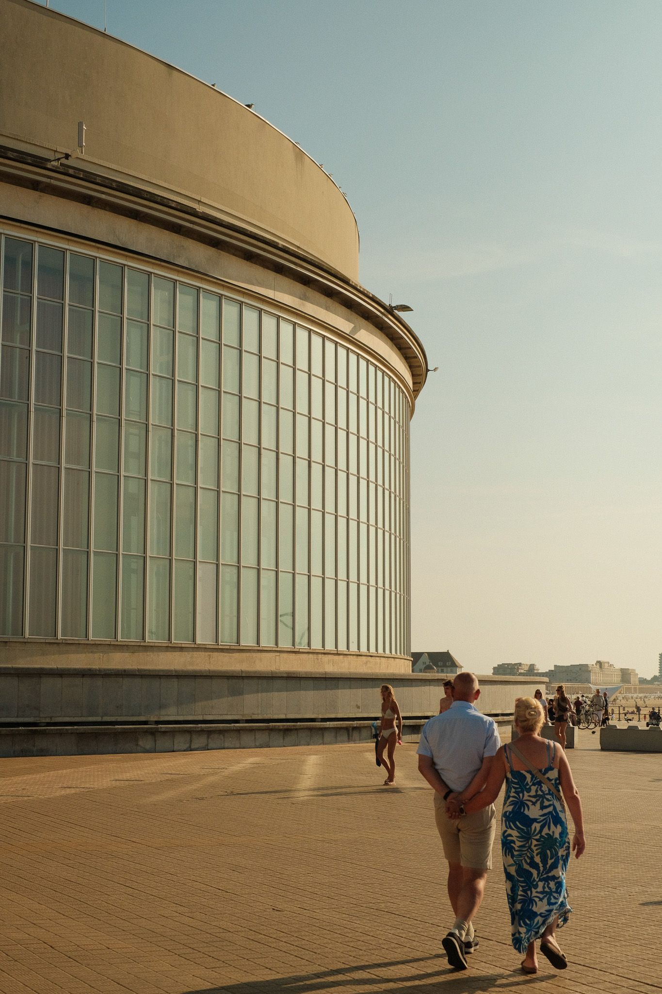 Modern curved building with glass windows and people walking on a sunlit terrace.
