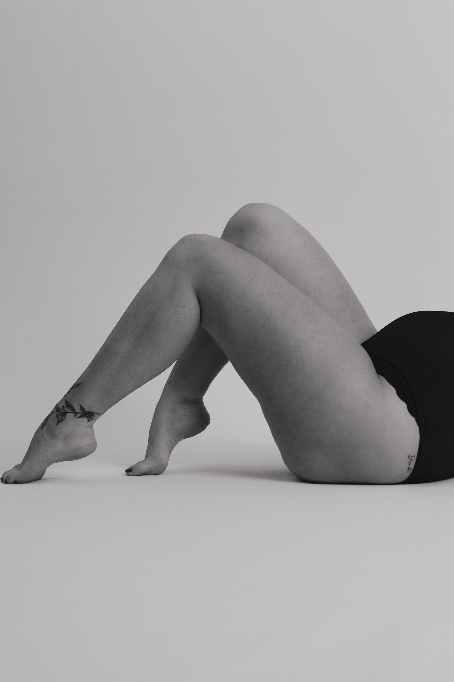 Black and white artistic nude study showing reclining figure with bent legs against neutral background.